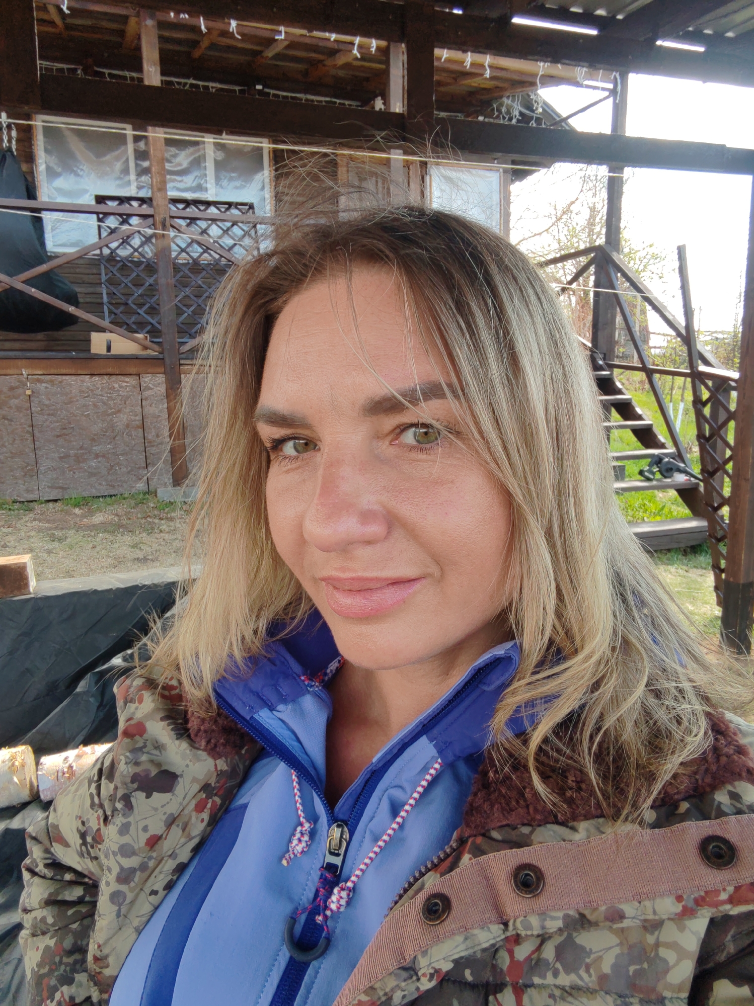 A woman with fair skin and blonde hair poses outdoors. She is wearing a camouflage jacket over a blue zippered shirt. The background shows a wooden building with stairs and windows. The location appears to be a rural or suburban setting, but information on whether it's the city of Koryaki, Russia, cannot be confirmed from this image.