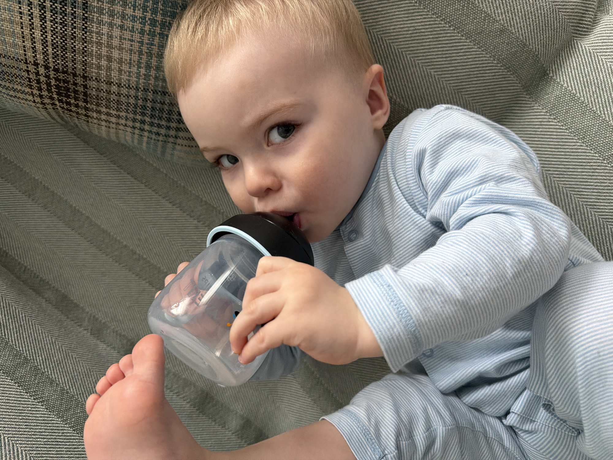 A baby with blond hair and light skin, wearing light blue striped pajamas, is lying on a couch. The baby is drinking from a clear plastic bottle with a black lid. The couch has a gray and beige textured fabric. The baby's foot is visible in the lower left of the frame. The city of Votkinsk, Russia, is not present or discernible in the image.