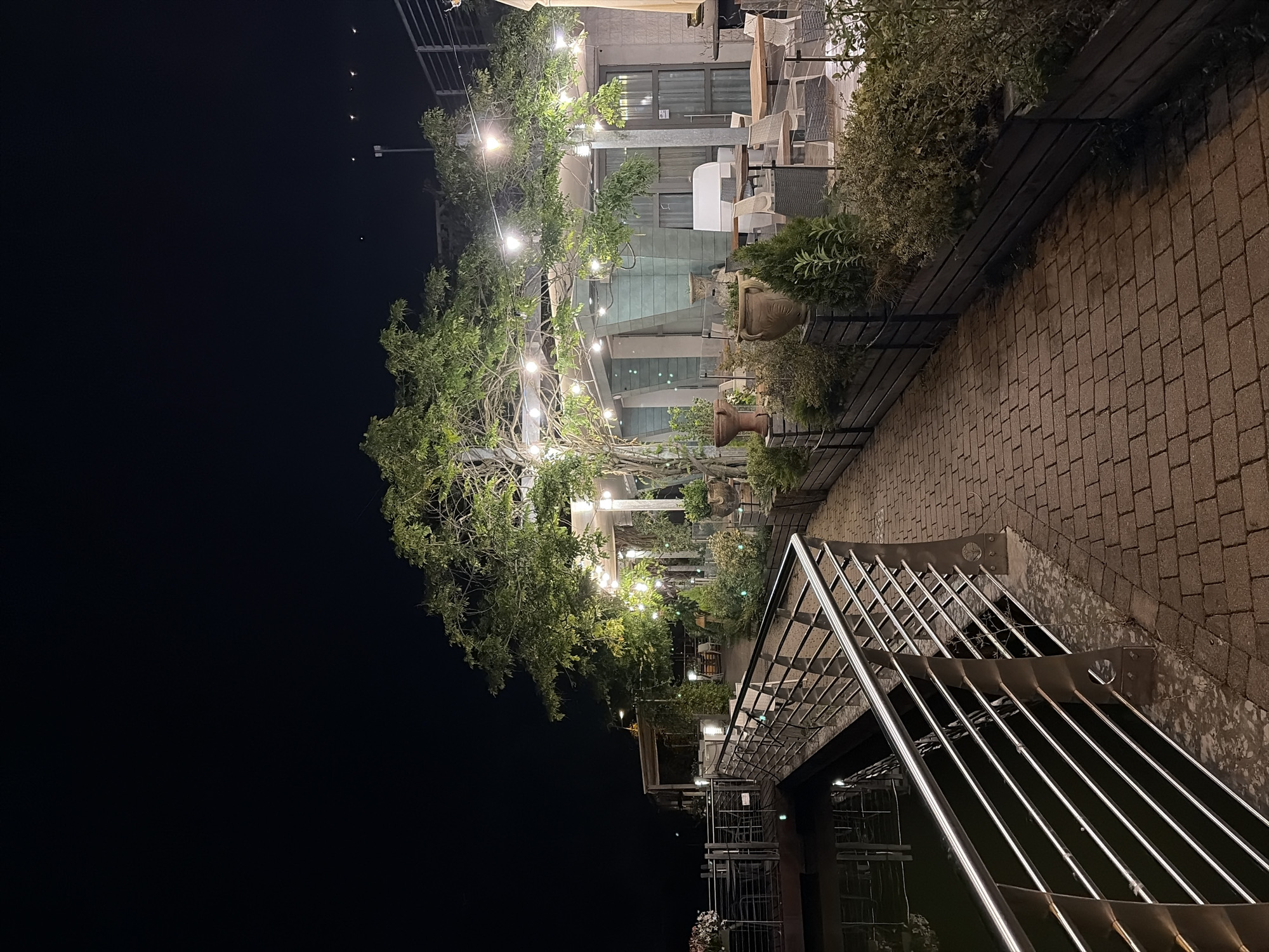 The image depicts an outdoor patio area at night, possibly a restaurant or cafe, situated near a body of water. A paved walkway, constructed from reddish-brown bricks, leads the viewer's eye through the scene. To the left of the walkway, a stainless steel railing borders the water, which appears dark and reflective.
The patio itself is illuminated by string lights draped across a large tree and a pergola-like structure. Several potted plants, including topiaries and larger decorative pots, are placed along the perimeter of the patio. Outdoor seating, consisting of tables and chairs, is visible in the background, suggesting it is set up for dining. A building with large windows and a light-colored facade forms the backdrop to the patio area. The overall ambiance is tranquil and inviting, with the warm glow of the lights contrasting with the dark night sky. The location in Trento, Italy, is not explicitly visible in the image but can be inferred from the prompt.