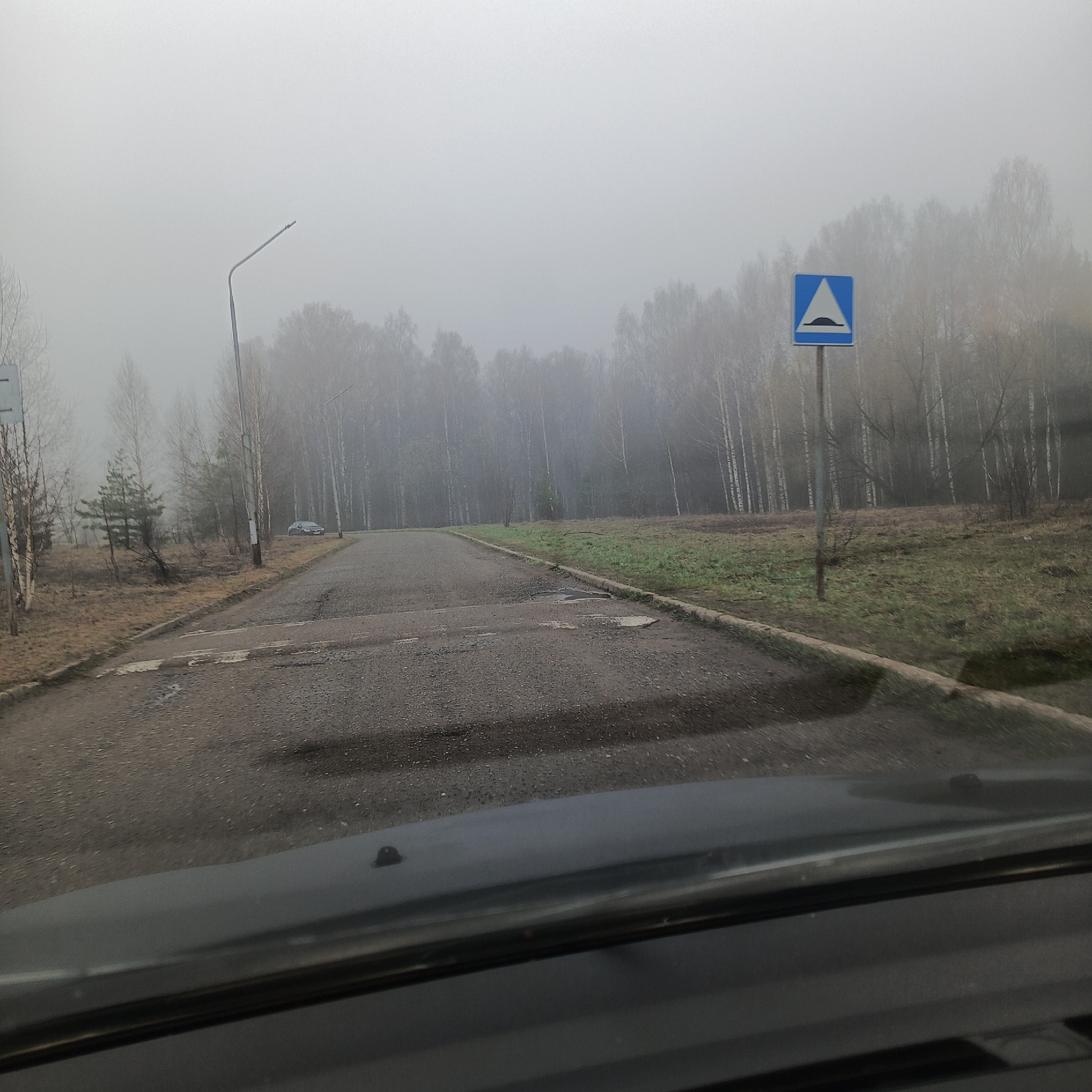 The image shows a road in Марковский, Russia, on a foggy day. The view is from inside a car, looking forward onto a straight, paved road with a speed bump. On the right side of the road is a traffic sign warning of the speed bump. Beyond the road and signs, a line of trees is visible through the fog. A lamppost is on the left side of the road.
