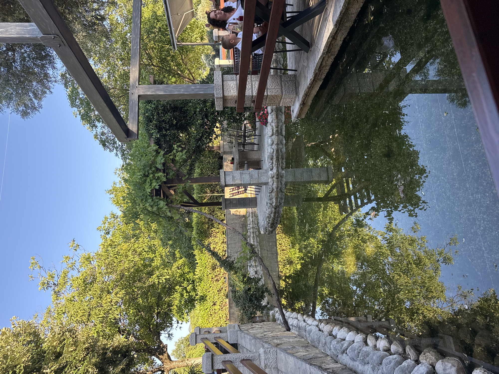 The image depicts an outdoor seating area adjacent to a shallow pond. In the foreground, a wooden table and benches are set up on a stone patio. A woman and a young boy are seated at the table, facing away from the camera. Behind them, a pergola structure with a dark wooden roof casts shadows. The pond, with its surface reflecting the surrounding trees and sky, is to the left of the seating area. A stone wall with a wooden railing lines the pond. In the background, lush green trees and hedges create a natural, verdant environment. The sky is clear and blue, suggesting a sunny day. The scene appears to be a restaurant or cafe with outdoor seating in a tranquil, garden-like setting.