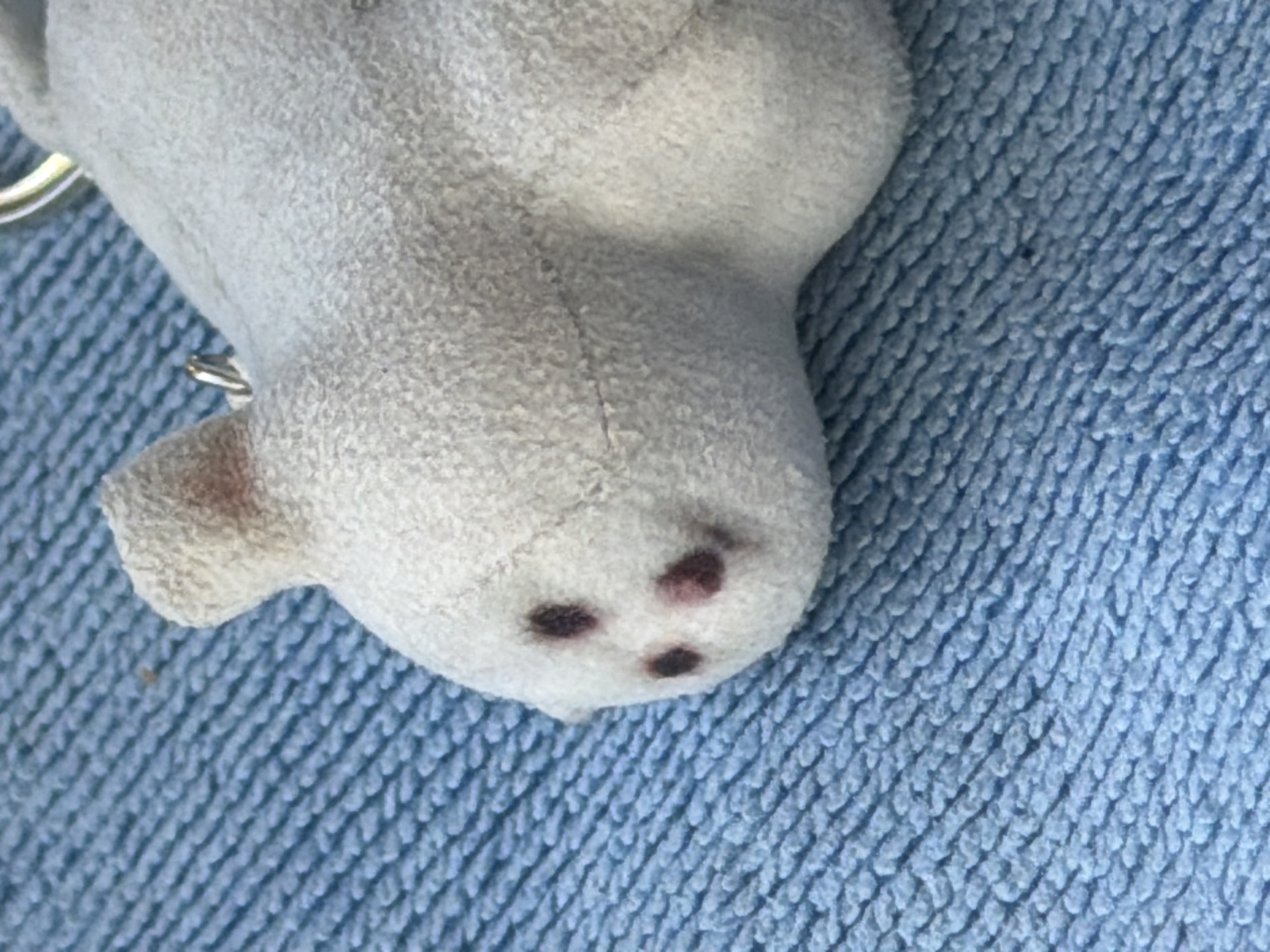 A close-up shot of a white plush toy with dark markings on its face rests on a blue, textured surface. The toy is positioned on the right side of the frame, with its head tilted to the left. Three dark, circular markings are visible on the toy's face, resembling eyes and a nose. A small metallic ring is attached to the top of the toy's head, suggesting it may be a keychain. The blue surface beneath the toy has a ribbed texture, possibly a towel or a blanket. The lighting is even, and the focus is sharp on the toy and its immediate surroundings. The background is out of focus, but the blue texture continues throughout. The image is devoid of any identifiable landmarks or contextual clues that would suggest a specific location within a city.