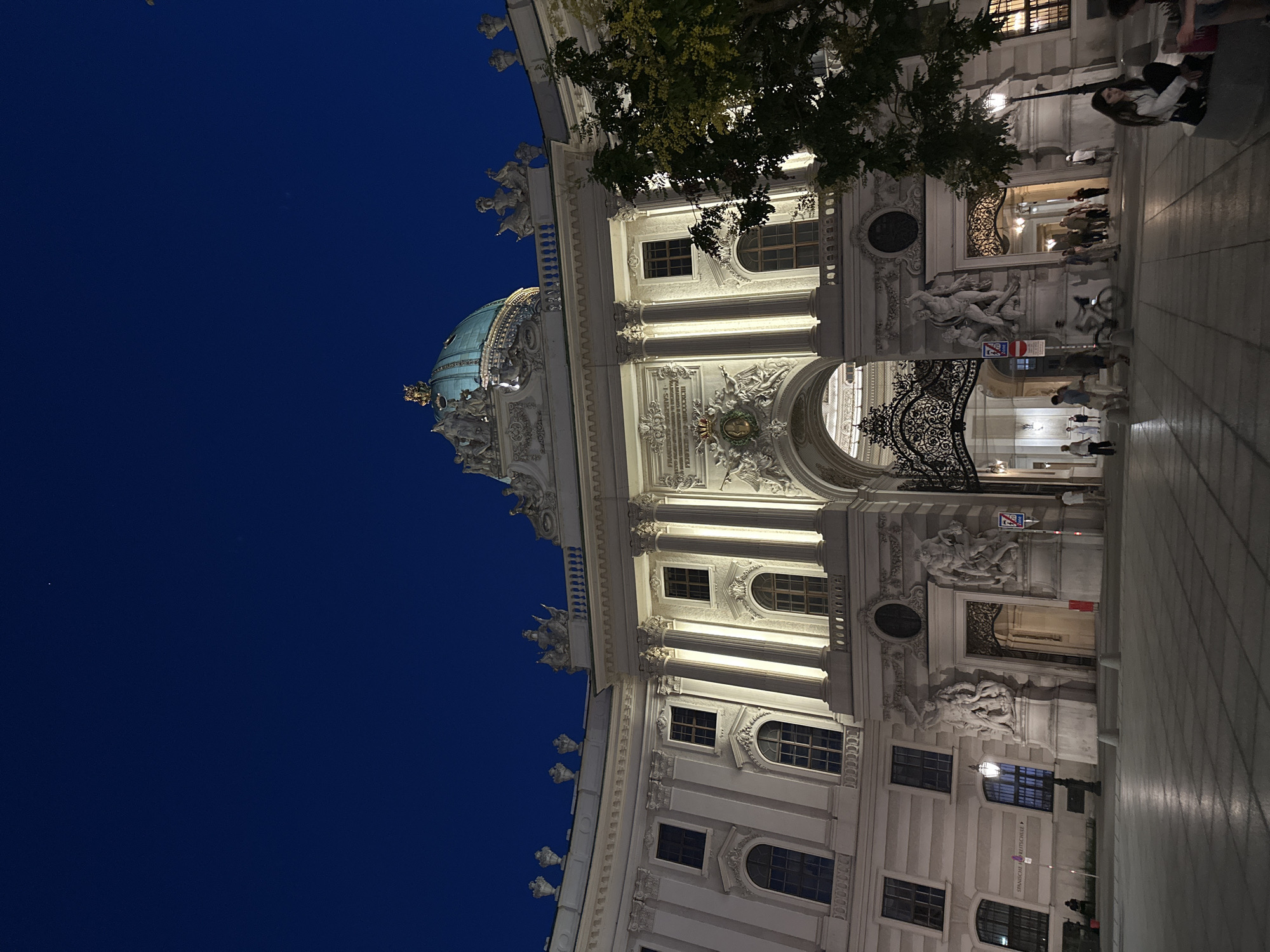 The image depicts a wide, curved building facade at dusk. The architecture is grand and ornate, featuring classical columns, intricate sculptures, and a prominent dome topped with a small spire. The building is illuminated by artificial lights, casting a warm glow on its pale stone exterior. A large, dark blue sky suggests it is either twilight or night.
In the foreground and along the pavement, several people are visible. They are casually strolling, with some gathered in groups. The pavement itself is tiled in a diagonal pattern, and a large, decorative wrought-iron gate stands open, suggesting an entrance.
To the left, a lush green tree with dark leaves is partially obscuring the building. To the right, a woman is seated on a low wall, looking towards the building. There is also a bicycle parked near the entrance and a few people walking in the background.
The overall impression is one of a peaceful evening scene in a historically significant and architecturally impressive location. The presence of people suggests this is a public space, possibly a landmark or a popular area for evening strolls.
Visible text includes "SPANISCHE REITSCHULE" on a sign next to a window, indicating the location is the Spanish Riding School in Vienna, Austria. There are also some directional signs with red and blue elements near the entrance.