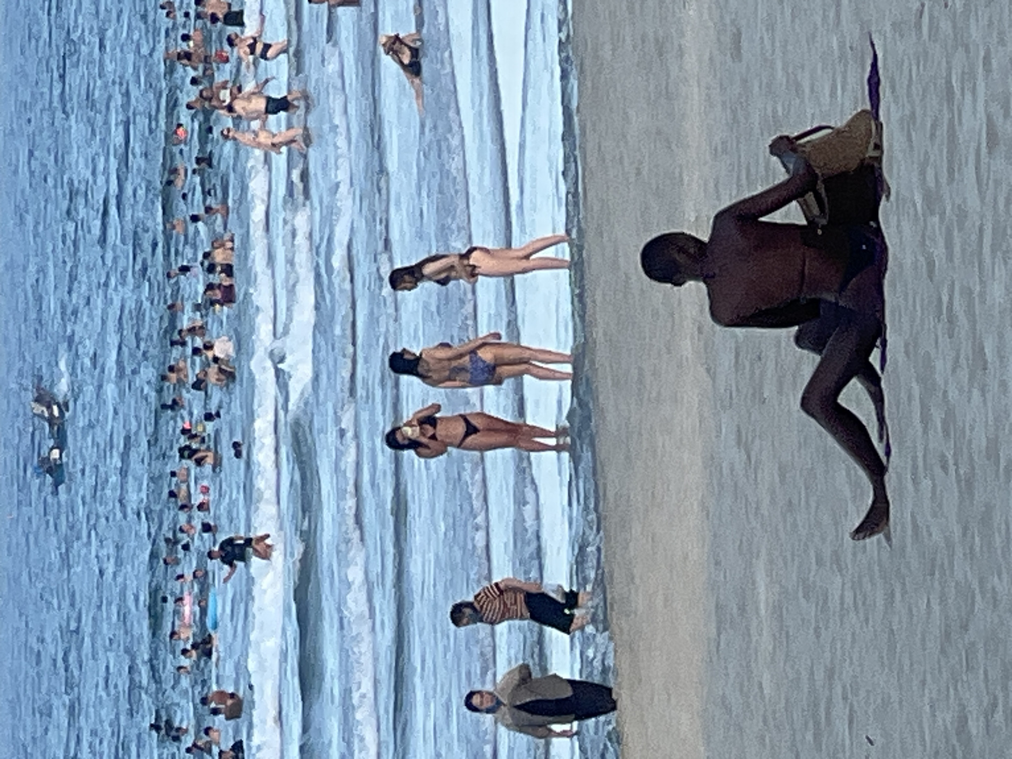 The image depicts a crowded beach scene, likely in Đà Nẵng, Vietnam, given the presence of many people swimming and wading in the ocean. In the foreground, a person with dark skin is sitting on the sand, facing away from the camera, wearing a dark bikini and with a bag next to them. Further into the water, several individuals are visible. Closer to the shore, two women in swimwear are standing, one on the left in a dark top and patterned bottoms, and another on the right in a blue bikini. To their left, a person in a striped shirt and dark shorts is partially submerged in the water. In the background, the ocean is filled with numerous people swimming, some with flotation devices. A boat is visible in the far distance on the water. The water is choppy with moderate waves. The overall atmosphere is that of a busy recreational area on a beach.
