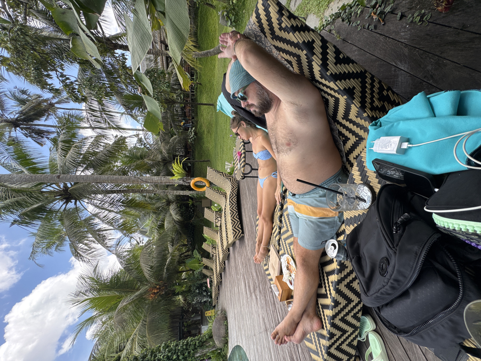 The image depicts a man and a woman relaxing at a resort in Peliatan, Indonesia. The man is lying down on a patterned woven chaise lounge, wearing a blue beanie, sunglasses, and swim trunks. Next to him is an empty glass with a straw, a can, and a black backpack. A charging phone and various footwear are also nearby. The woman is lying next to him in a blue bikini. The resort appears to be situated amidst lush tropical vegetation including palm trees under a partly cloudy sky. Additional chaise lounges are seen lined up in the distance.