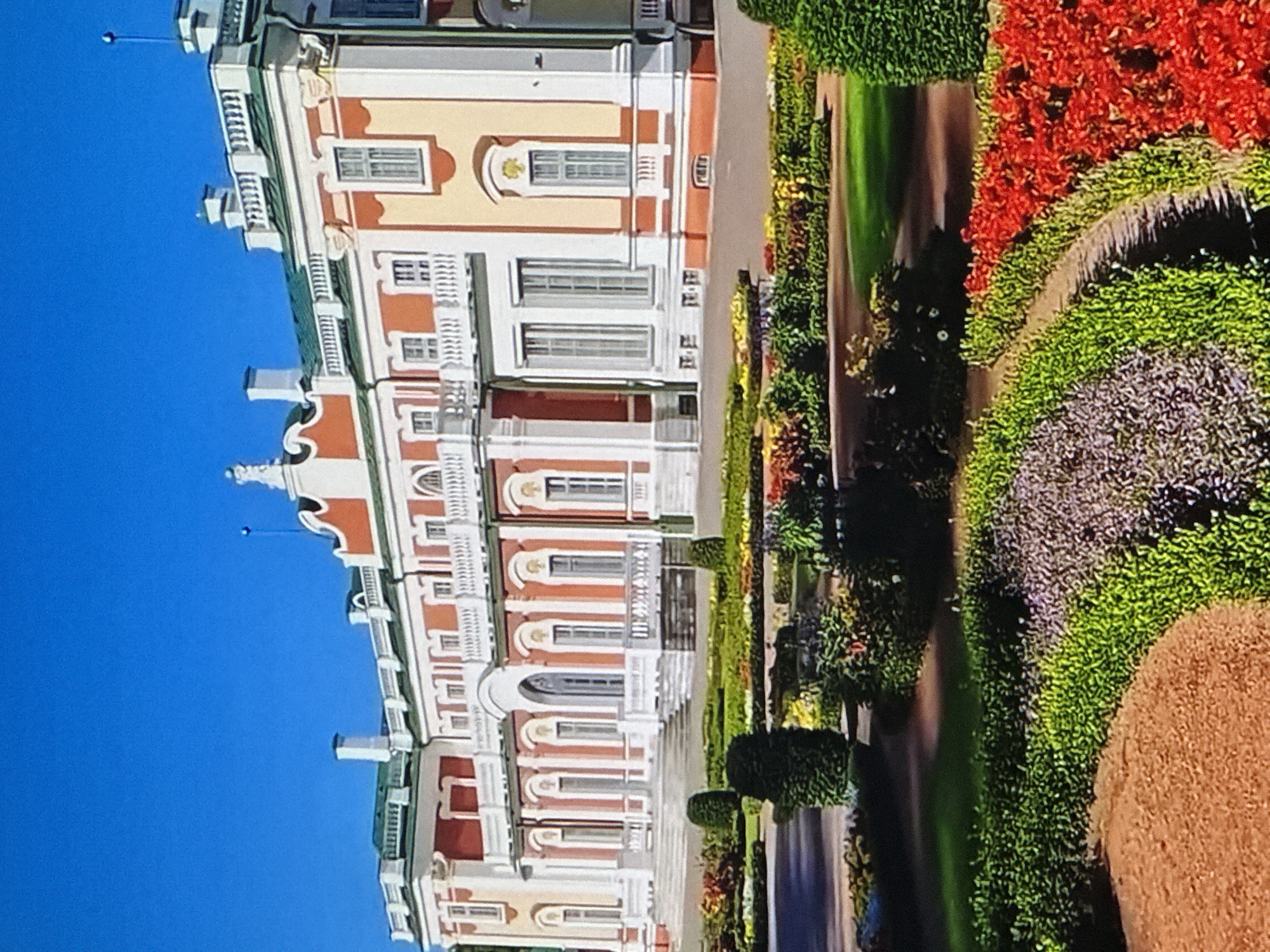 The image depicts the Kadriorg Palace in Tallinn, Estonia. The palace is a grand baroque building with a red and yellow facade, white columns, and a green roof. It is set against a clear blue sky. In front of the palace, there is a formal garden with manicured bushes, colorful flower beds, and pathways. The garden features a variety of plants, including red, purple, and green foliage. The scene is well-lit, suggesting it was taken on a sunny day. There are no people visible in the image.