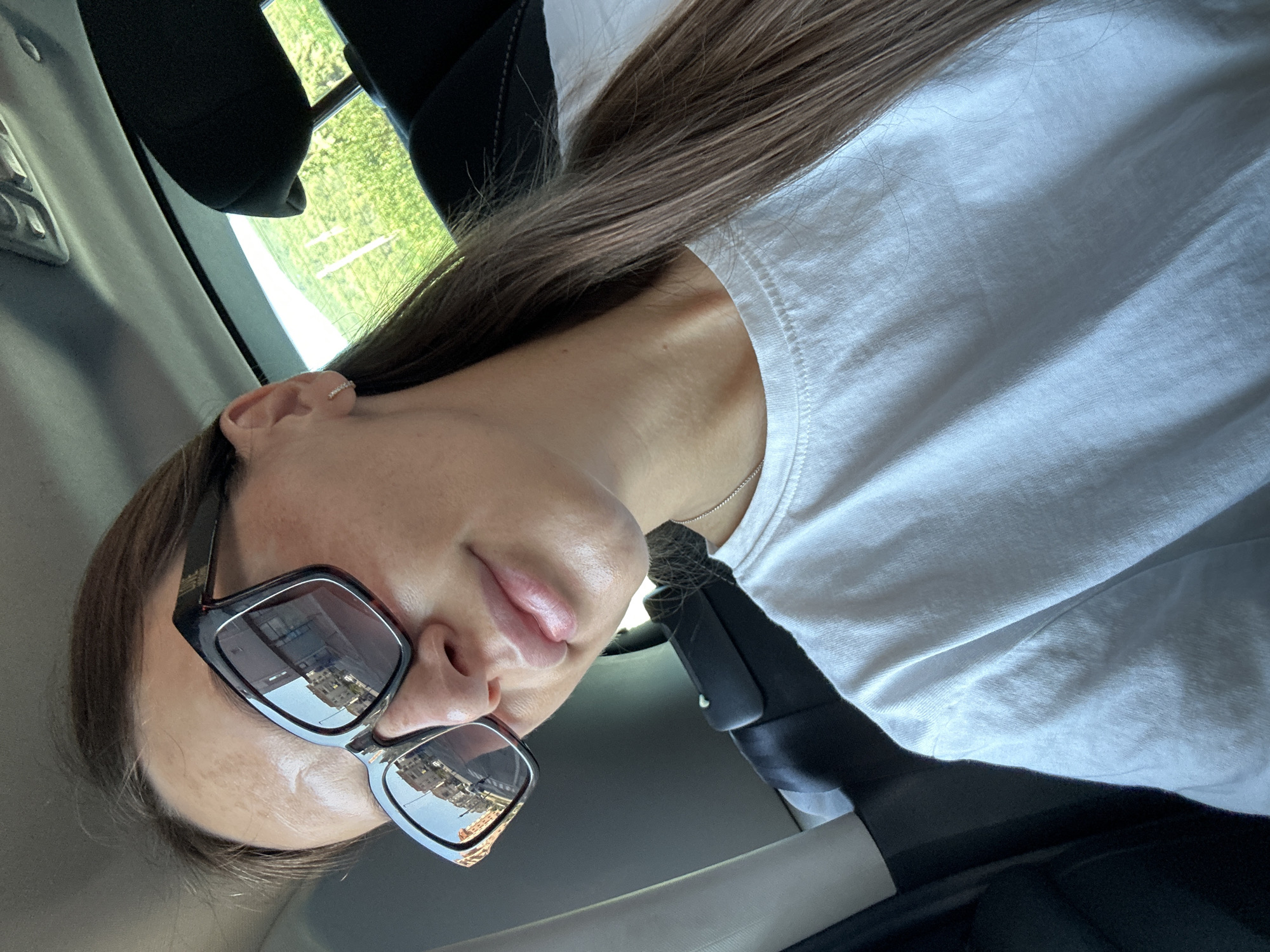 A young woman is captured in a close-up selfie taken from within a vehicle, likely in the city of Donji Pobori, Montenegro. She is wearing large, dark-rimmed sunglasses that reflect buildings, suggesting an urban or suburban environment. The woman has long, brown hair that extends past her shoulders. She is also wearing a simple white t-shirt and a thin silver necklace. A seatbelt is visible across her chest, indicating she is the passenger. The background is blurred, but hints of green foliage and the interior of a car, including a headrest, are discernible.