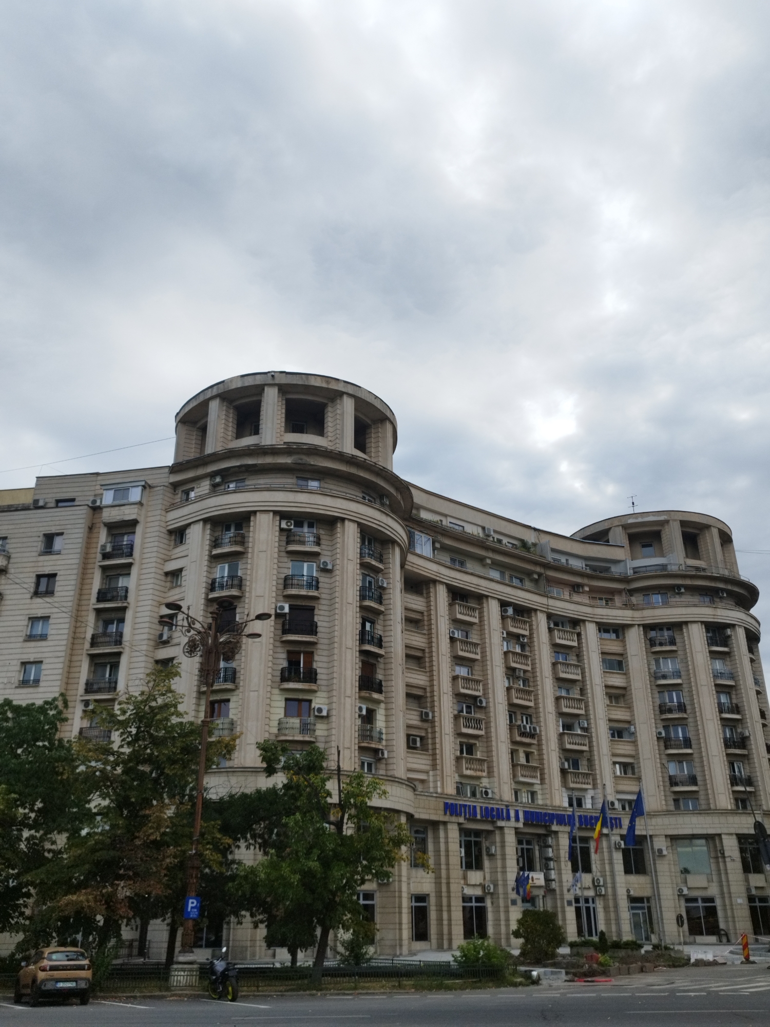 The image shows a large, light-colored building in Bucharest, Romania, with a semicircular facade. The building has many floors, each with numerous windows and balconies. Some of the windows are open, and air conditioning units are visible on the exterior walls. There are several flags displayed on the building's facade, including the Romanian flag and the European Union flag. Blue lettering above the ground floor windows reads "POLITIA LOCALA A MUNICIPIULUI BUCURESTI", indicating it is the Local Police headquarters for Bucharest. The building is surrounded by trees and a street with a parked car and motorcycle. The sky is overcast with heavy clouds.