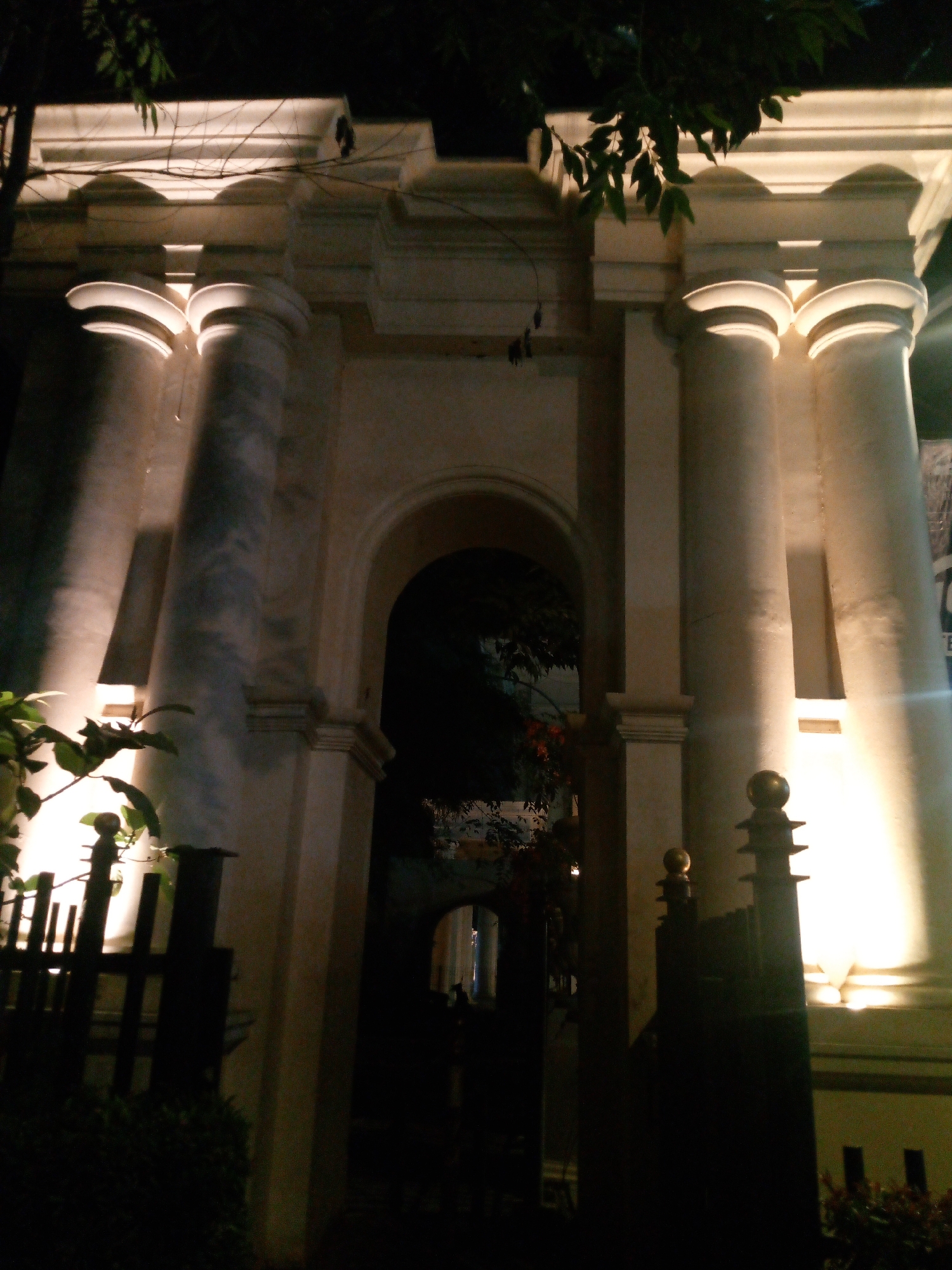 The image shows the exterior of a building at night, illuminated by artificial lighting. The architecture features large, fluted columns and an arched entryway. A dark, ornate fence or gate is in the foreground on the left, with foliage obscuring part of it. On the right, another dark structure, possibly a gate or fence with finials, is visible. The lighting creates dramatic shadows and highlights on the building's facade. The scene is located in the city of Bitung Jaya, Indonesia. There are no people visible in the image.
