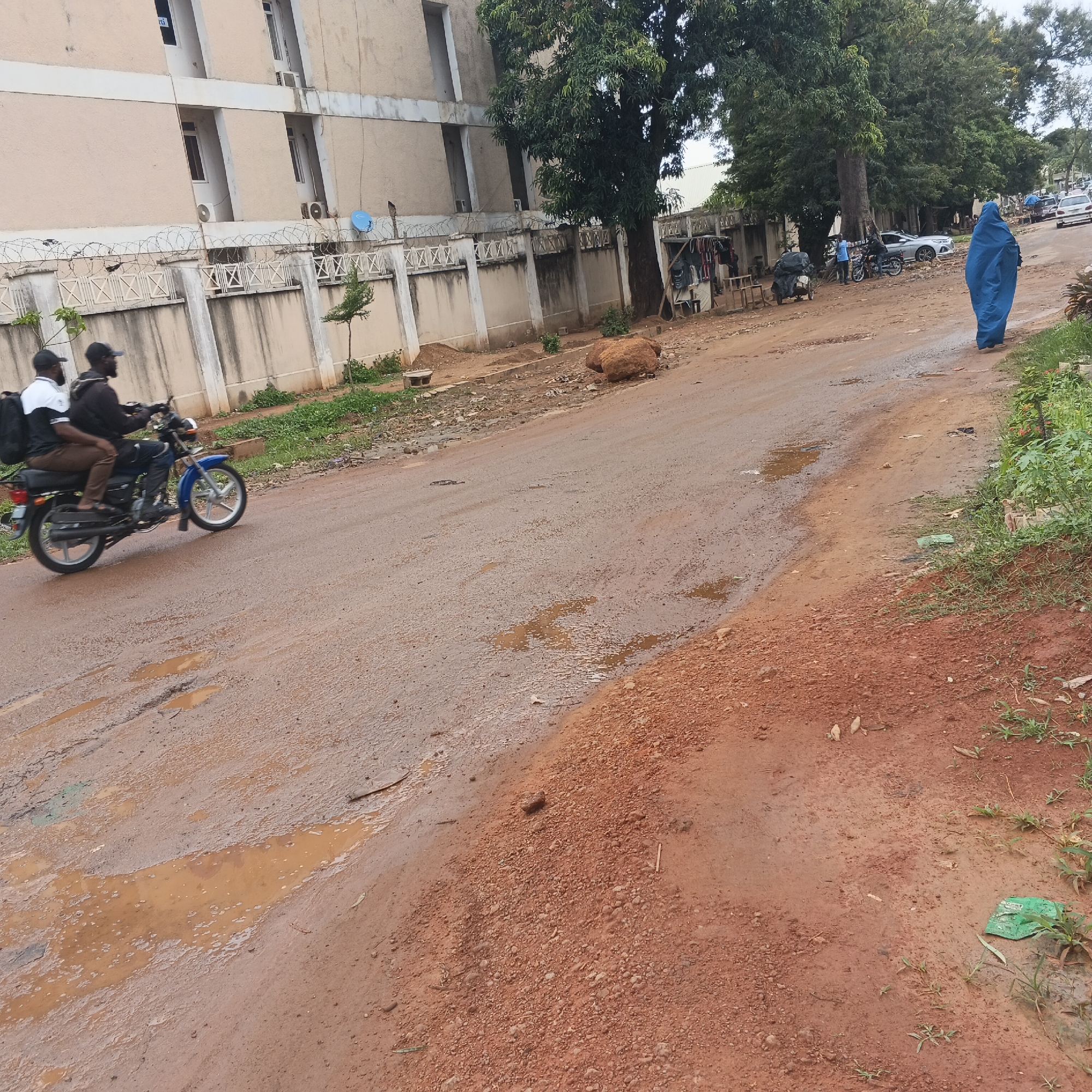 The image depicts a dirt road in Shaba, Nigeria, with a section of a light-colored, multi-story building visible in the background. The road is unpaved and shows signs of wear, with several puddles of water scattered across its surface. A fence with barbed wire runs along the side of the building. Two individuals are riding on a blue motorcycle in the foreground, moving from left to right. The rider wears a dark hoodie and a black cap, while the passenger behind him wears a white and black top and a black cap. A large black bag is secured to the back of the motorcycle. Further down the road, a person in a blue covering is walking away from the camera. Several cars are parked or driving in the distance. The environment appears to be an urban or semi-urban area, with vegetation growing along the side of the road. The ground is a reddish-brown, characteristic of laterite soil.