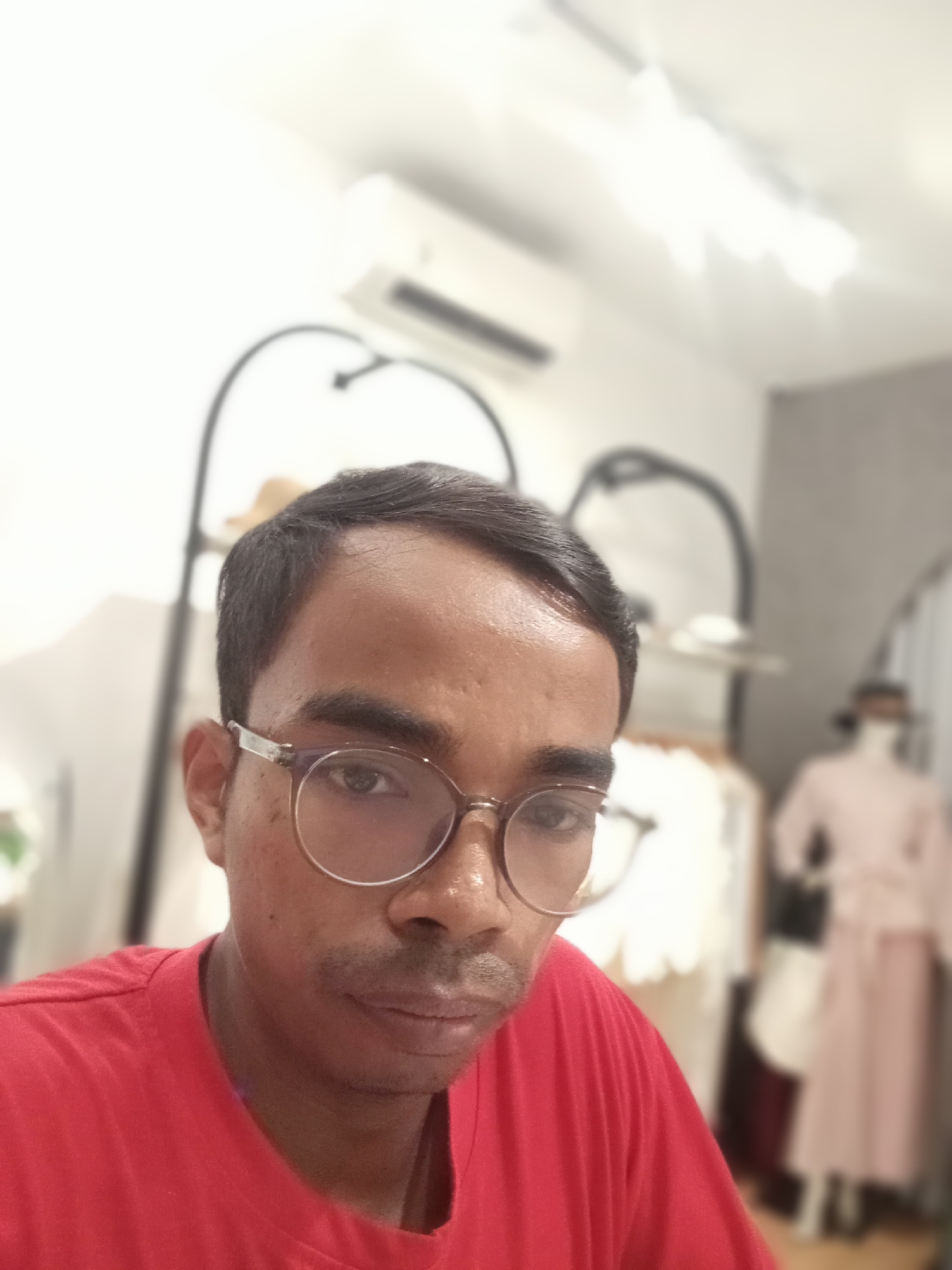 A medium-shot, selfie-style photograph captures a man with dark hair and glasses, wearing a red t-shirt. He is positioned in the foreground, with his face angled slightly towards the camera. His gaze is directed at the lens. The background is softly blurred, revealing what appears to be an indoor setting, possibly a clothing store or studio, with clothing racks and mannequins visible. The lighting is bright, suggesting an indoor environment. The location is specified as Larangan, Indonesia.