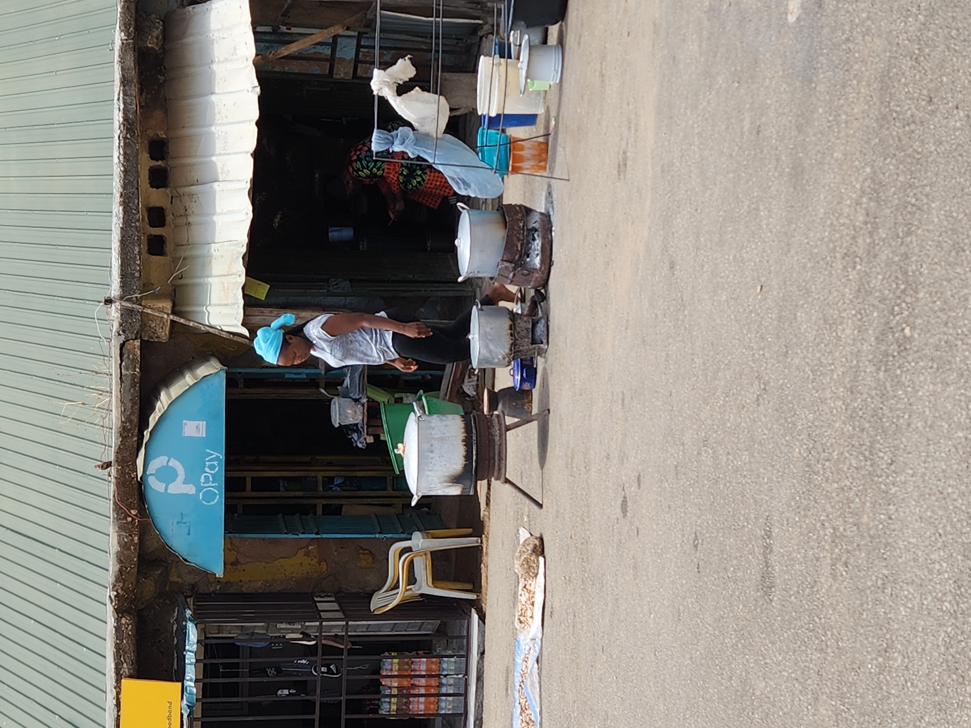 The image shows a street scene in Kabong, Nigeria. A woman wearing a blue headscarf and a white t-shirt is standing near several large cooking pots set on open fires, likely preparing food for sale. Behind her is a building with a corrugated metal awning and a blue "Opay" sign. Inside the building, other people are visible, one wearing a colorful patterned dress. Hanging above the pots are various containers. On the ground near the cooking area, there is what appears to be a bag of food items and plastic chairs. A variety of items are for sale on the shelves in the building. The street is paved, and there are no vehicles visible.