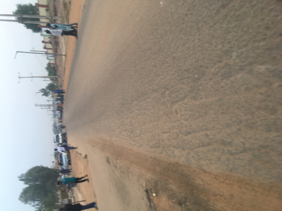 A long, gray asphalt road stretches into the distance. People walk along the side, and cars and motorcycles are parked or moving in the same direction. Utility poles line the side of the road with trees scattered throughout. The sky appears overcast and hazy. The road surface has some texture and wear.