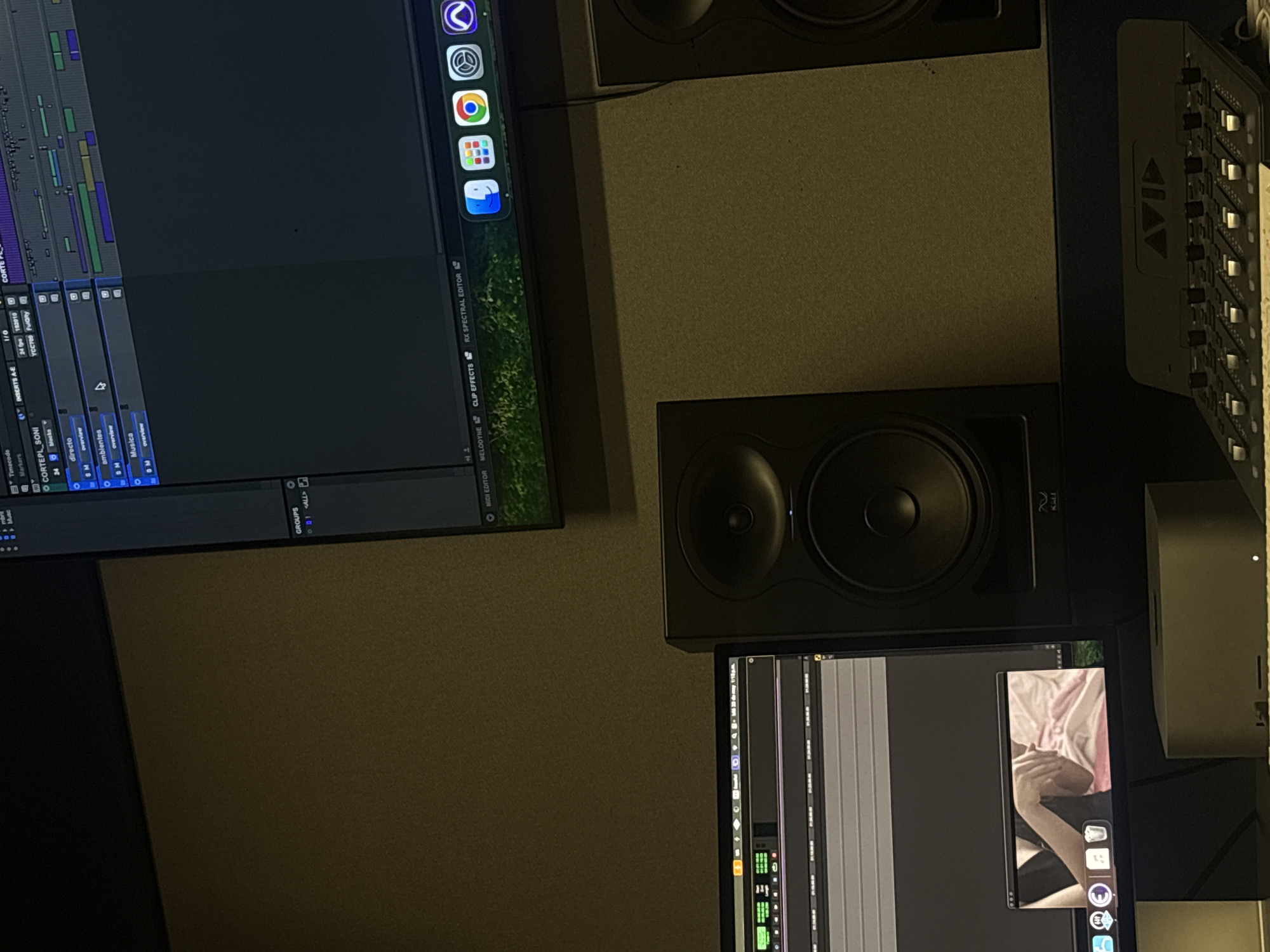 The image shows a recording studio setup. On the left, a computer monitor displays digital audio workstation (DAW) software with audio tracks and editing tools. The icons on the screen suggest the use of Apple software. Above the monitor, a studio monitor speaker is visible. To the right, an Avid control surface is partially visible along with a second studio monitor speaker below. A second computer monitor can be seen below the studio monitor. The recording studio likely has acoustic treatment on the walls, as evidenced by the textured panel behind the monitors. The setting of the image could not be derived from the available image data.