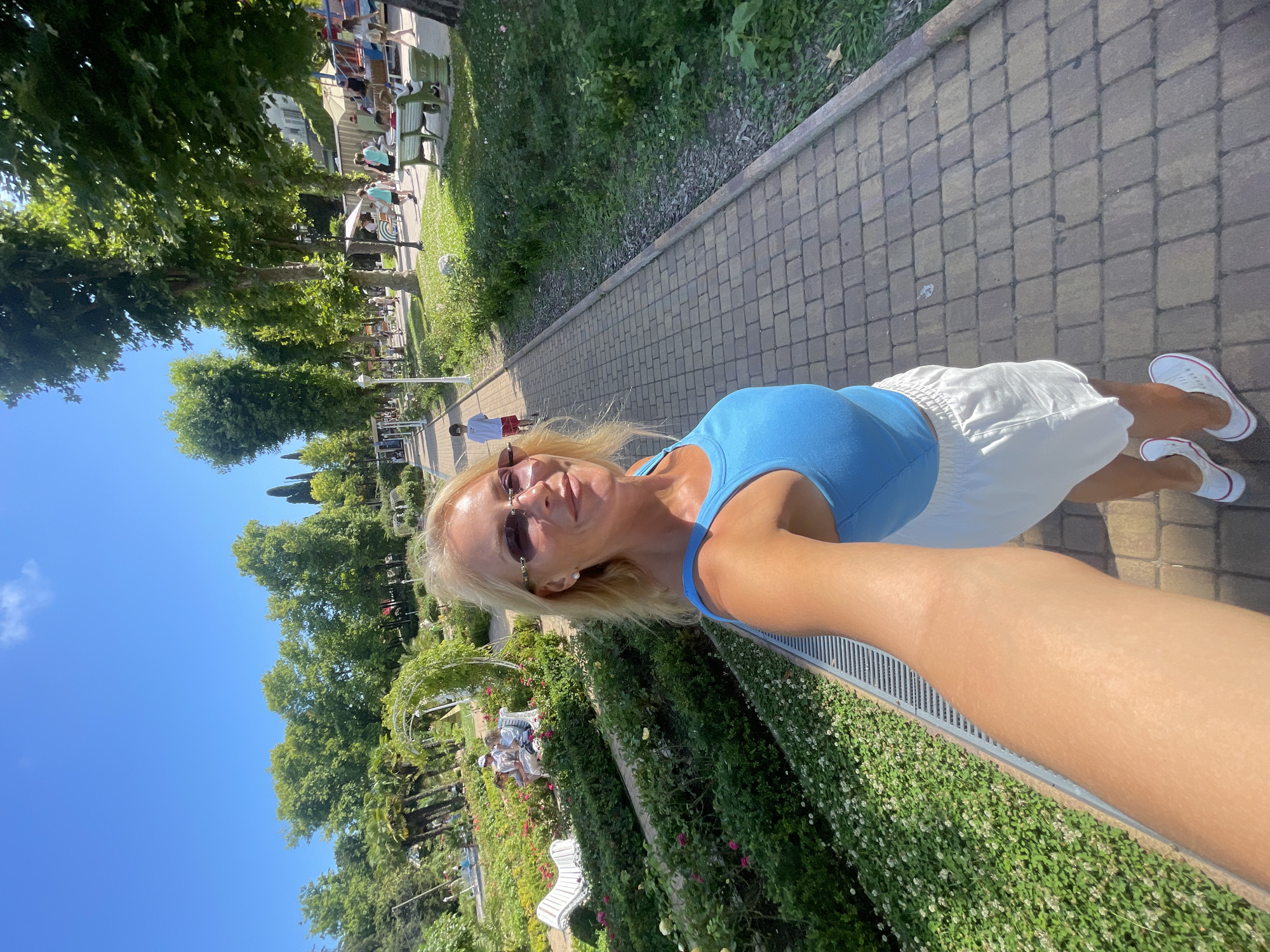A blonde woman in sunglasses, a blue tank top, and white shorts takes a selfie with her left arm extended in a park on a sunny day. She is walking on a paved walkway lined with green bushes and flowers. In the background, a young boy in a white shirt and red shorts walks away from the camera, and several people are visible sitting on benches and near a playground. The location is identified as Sochi, Russia.