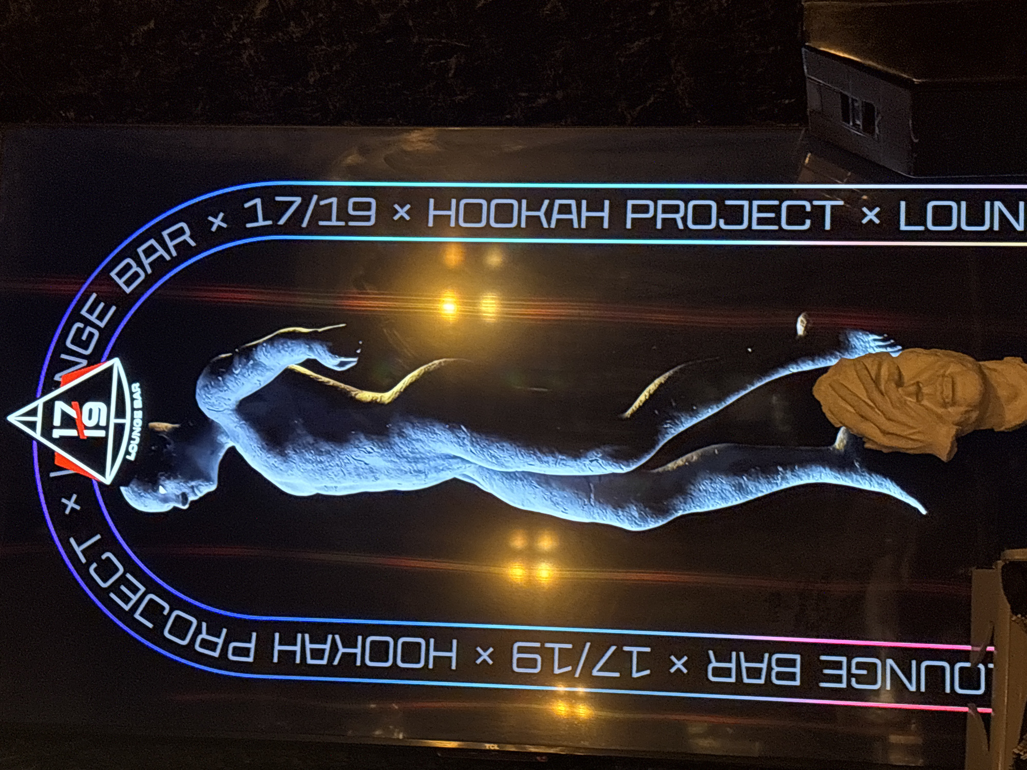 An exterior shot shows a neon sign arching over a blue-toned statue of a person, with the words "LOUNGE BAR" and "17/19 HOOKAH PROJECT" displayed vertically along the sides of the arch. A triangular logo with the numbers "17" and "19" is visible at the apex of the arch. Behind the statue, the blurry lights of a city street are visible, with the city identified as Kazan, Russia. The statue itself is of a nude figure, and a second, disembodied head sculpture is positioned at the base of the statue, obscuring its feet. The statue is lit from below, casting a blue glow. The neon sign is illuminated with pink and blue lights.