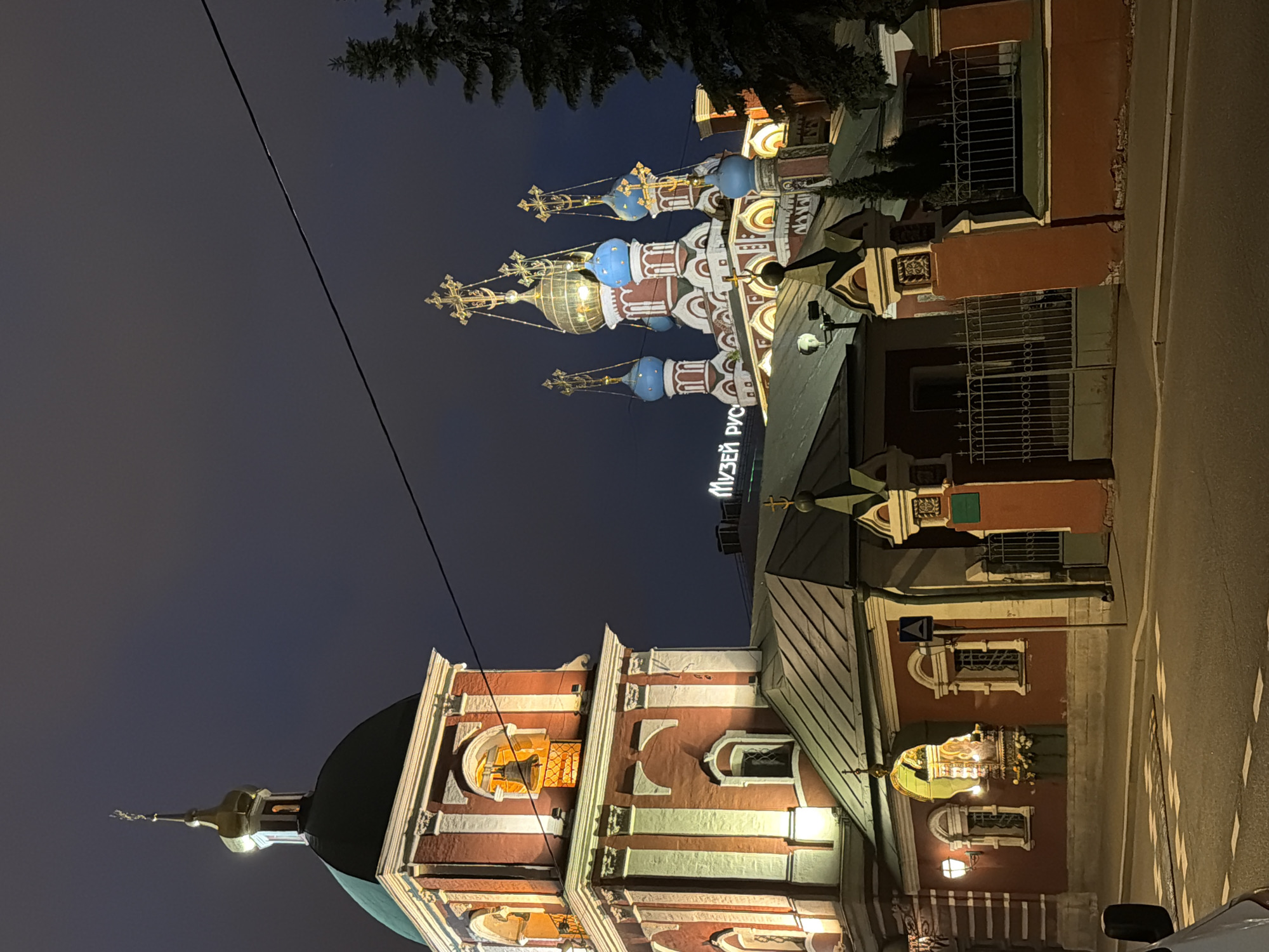 Captured at night, the image showcases a multi-tiered Russian Orthodox church illuminated against a dark sky. The church exhibits a traditional design with red and white masonry, arched windows, and multiple domes topped with golden crosses. The domes vary in color, including blue, silver, and gold. Part of the church bears a sign in Cyrillic script. Adjacent to the church is a road with painted zebra stripes and sidewalks. The scene is in Moscow, Russia.
