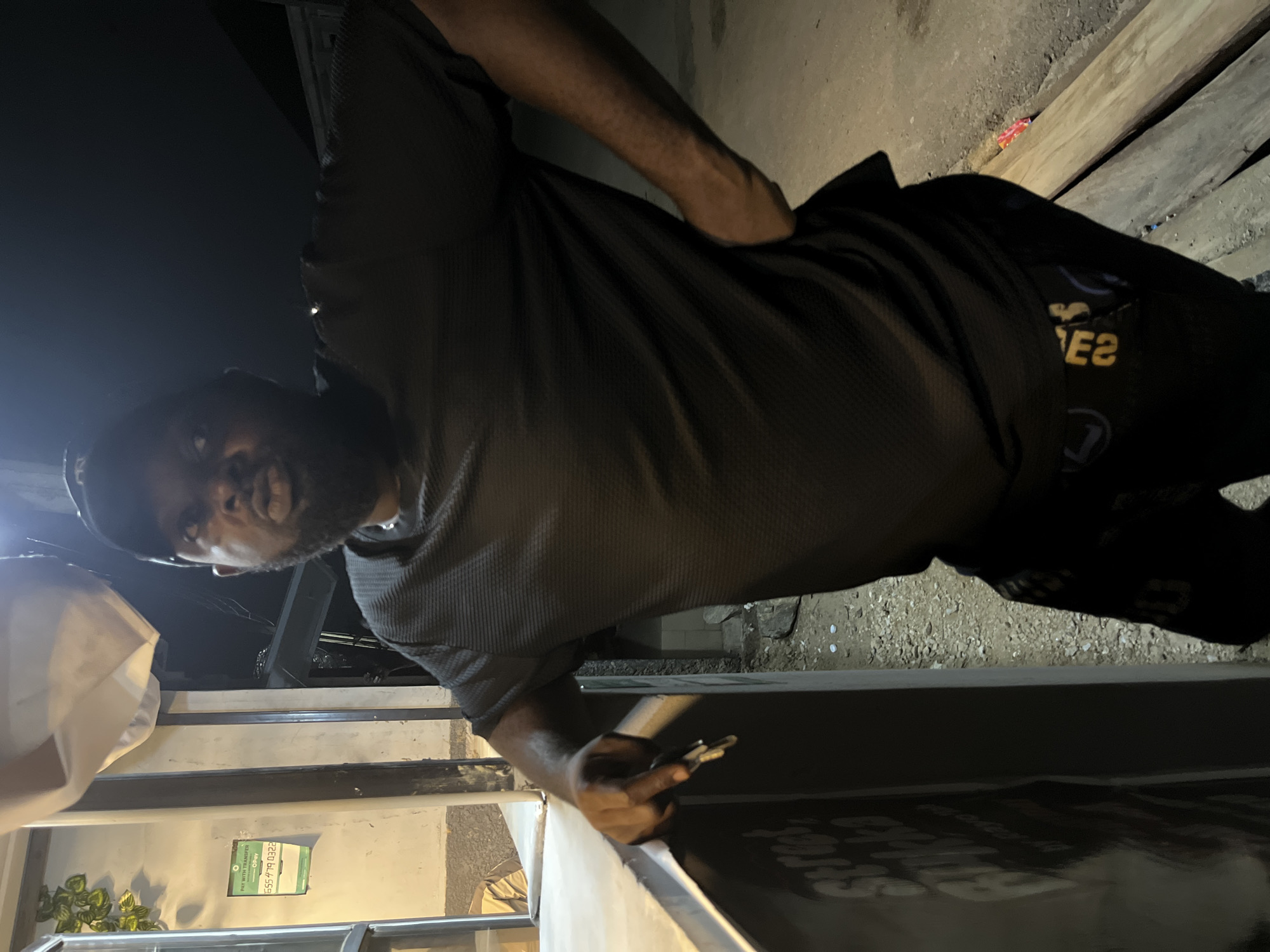 A man is standing near a structure, possibly a counter, in an outdoor area. The environment appears to be a street setting with mixed surfaces and lighting. The man is holding a small object. The man is wearing a black shirt and a hat. The location is Kubwa, Nigeria.