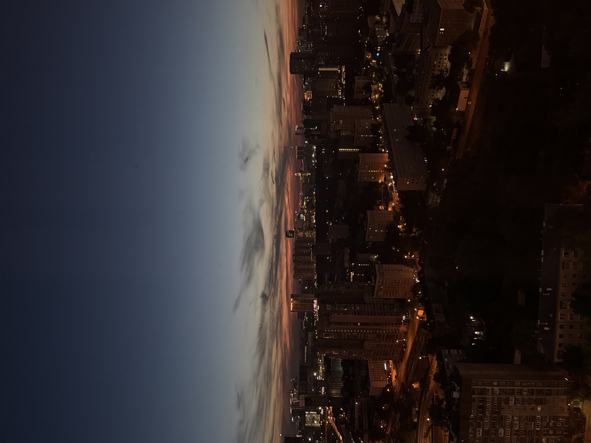 This media file captures a panoramic view of a city transitioning from twilight to night, with the image rotated 90 degrees clockwise. The left side of the frame features the sky, displaying a gradient of colors from a deep, dark blue at the far left to warmer oranges, pinks, and purples towards the center. Elongated, wispy clouds streak across this colorful horizon.
The right side of the image reveals a sprawling urban landscape, largely enveloped in darkness. Numerous buildings, appearing as multi-story structures and taller high-rises, are visible, with countless windows casting yellow and white light. Streetlights illuminate roads and paths, creating patterns of light that snake through the city. Darker, unlit areas, possibly parks or undeveloped land, are interspersed between clusters of illuminated buildings. The perspective suggests a high vantage point, looking down over the expansive city. No specific human activity or people are discernible, and there is no visible text. The overall impression is one of a vast, tranquil city settling into the night.