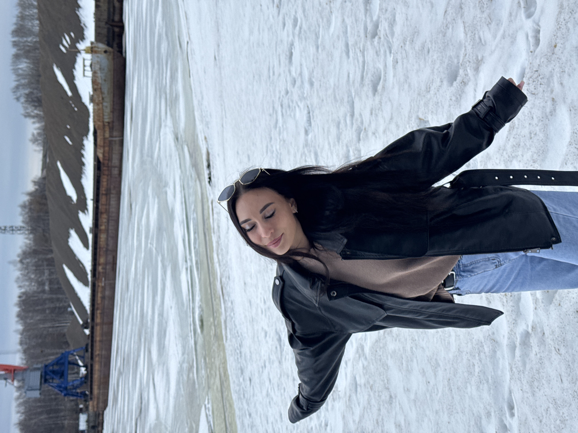 A woman with long, dark hair wearing sunglasses and a black leather jacket over a light brown sweater and blue jeans stands outdoors in a snowy environment. The woman's eyes are closed, and she appears to be smiling slightly. Behind her, there is a landscape dominated by snow and ice, along with industrial infrastructure, including a structure that resembles a long building and a blue crane. The location is Chaikovsky, Russia.
