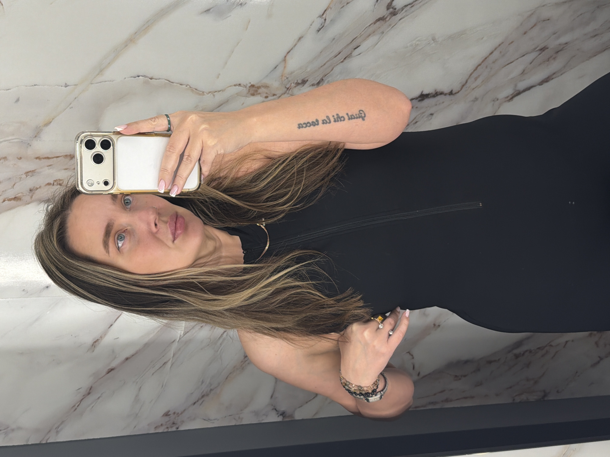 A woman takes a selfie in what appears to be a bathroom with marble walls. She is holding a gold iPhone in her left hand and is wearing a black dress. She has brown hair with blonde highlights and is wearing multiple rings and bracelets. The location is unknown.