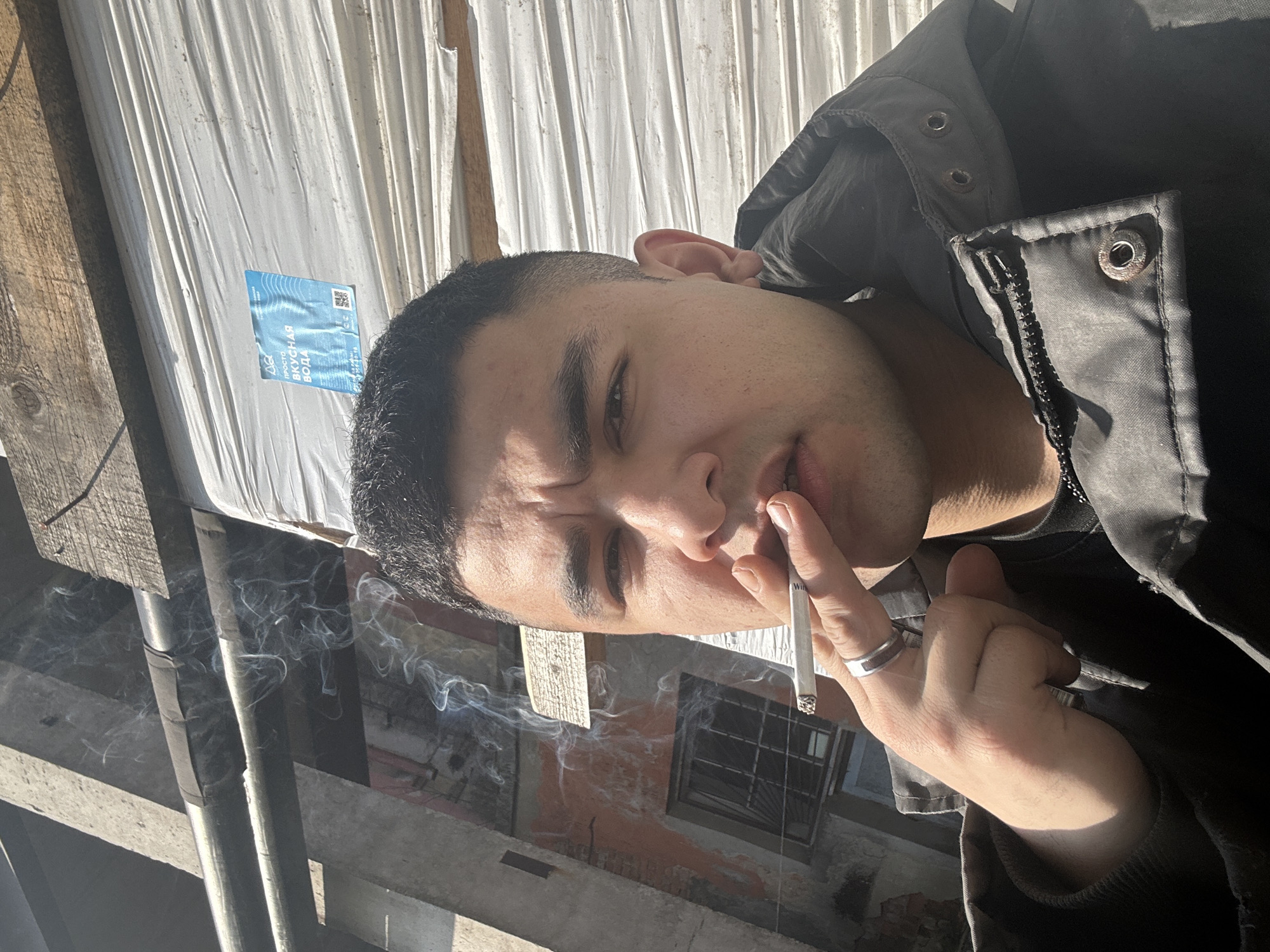 A man with short dark hair and light skin is shown in a close-up, low-angle shot. He is holding a lit cigarette in his right hand, close to his lips. He wears a dark jacket. Smoke from the cigarette is visible, swirling in the lower left area of the frame. The background includes a white tarp, wooden beams, and what appears to be an exterior urban setting with faded brick walls and window. A small blue advertisement is affixed to the tarp. The photo was taken in the city of Fornosovo, Russia.