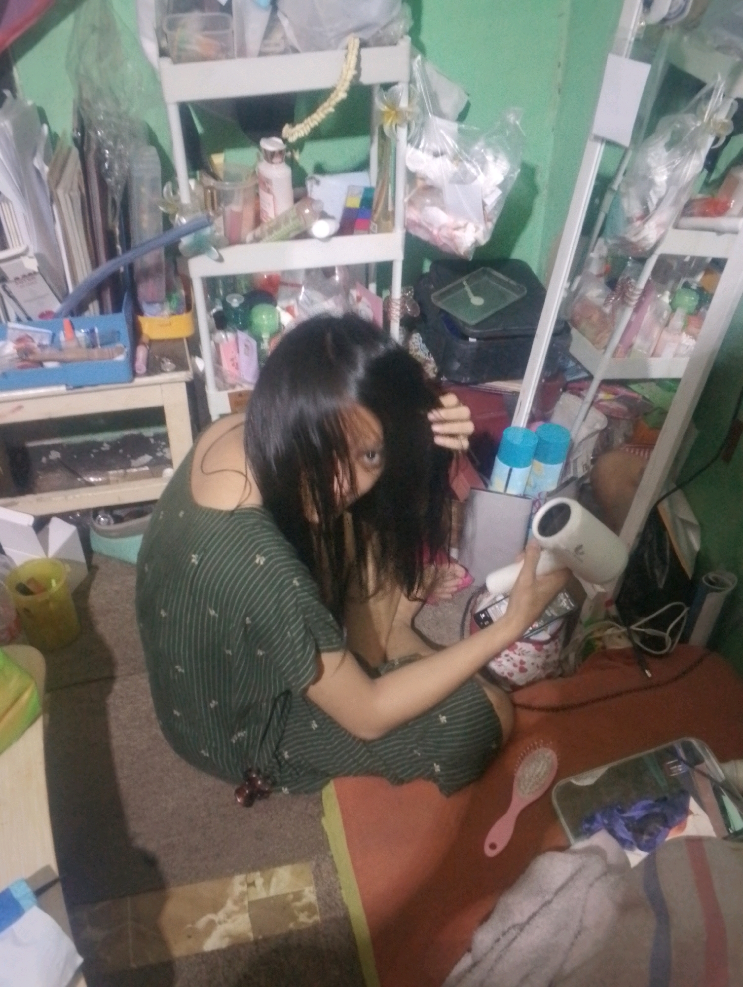 The image depicts a person in what appears to be a cluttered bedroom or living space in Depok, Indonesia. The person, with long dark hair, is wearing a green and white striped garment and is holding a hairdryer. Their hair is wet, suggesting they may have recently washed it. A pink hairbrush is visible on a reddish-brown surface in the foreground. In the background, shelves are filled with various items, including bottles, containers, and what seem to be personal belongings. The room is dimly lit and filled with objects, creating a sense of disarray. The overall impression is of a candid moment captured in a personal, everyday setting within an urban environment.