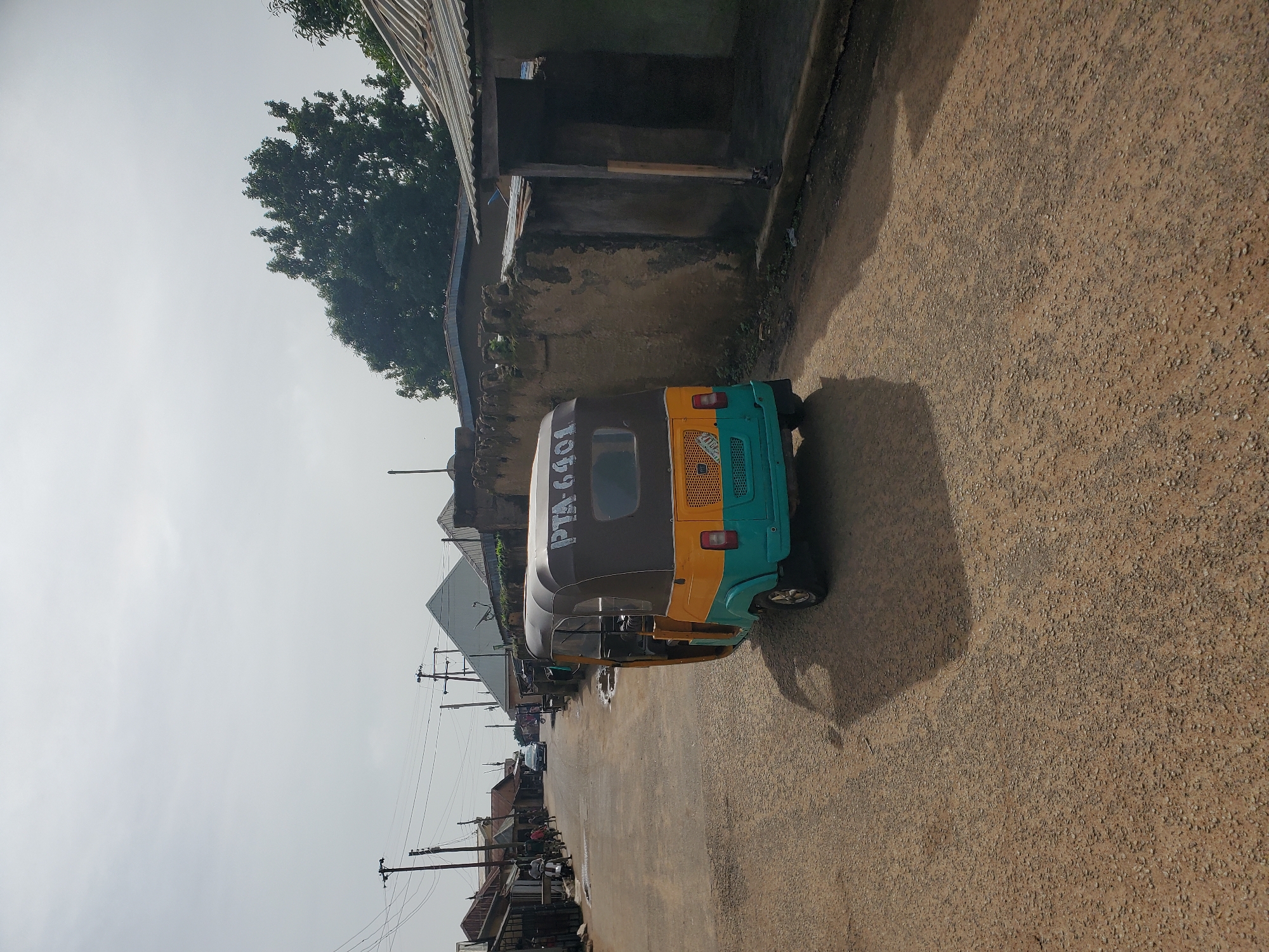 In Jos, Nigeria, an open-air street scene unfolds under a hazy sky. A yellow and turquoise auto-rickshaw, marked with "PTA 6401", dominates the foreground, parked on a gravelly, uneven road. The vehicle, a common mode of transport in the region, displays wear and tear consistent with its use.
To the right of the auto-rickshaw, a weathered, mud-brick wall encloses a compound with a corrugated metal roof. The structure appears aged, with cracks and signs of deterioration in the wall. Further down the street, other buildings line both sides, their roofs predominantly made of corrugated metal. Some structures appear to be shops or residences, with open doorways and windows suggesting commercial or domestic activity.
On the left side of the road, a few individuals are visible. One person stands near the entrance of a building, while others are seen further down the street, some possibly on motorcycles or bicycles. A white car is seen moving away from the viewer in the distance, indicating ongoing traffic. Power lines with utility poles crisscross the sky, a common feature in urban and semi-urban environments. The overall impression is of a typical day in a developing city, with elements of both daily life and the challenges of infrastructure maintenance. The ground is a mix of gravel and dirt, with some puddles indicating recent rain or poor drainage. A large tree with dense foliage stands behind the buildings on the right, offering a stark contrast to the man-made structures. The environment suggests a warm climate, with the hazy sky possibly indicating dust or humidity.