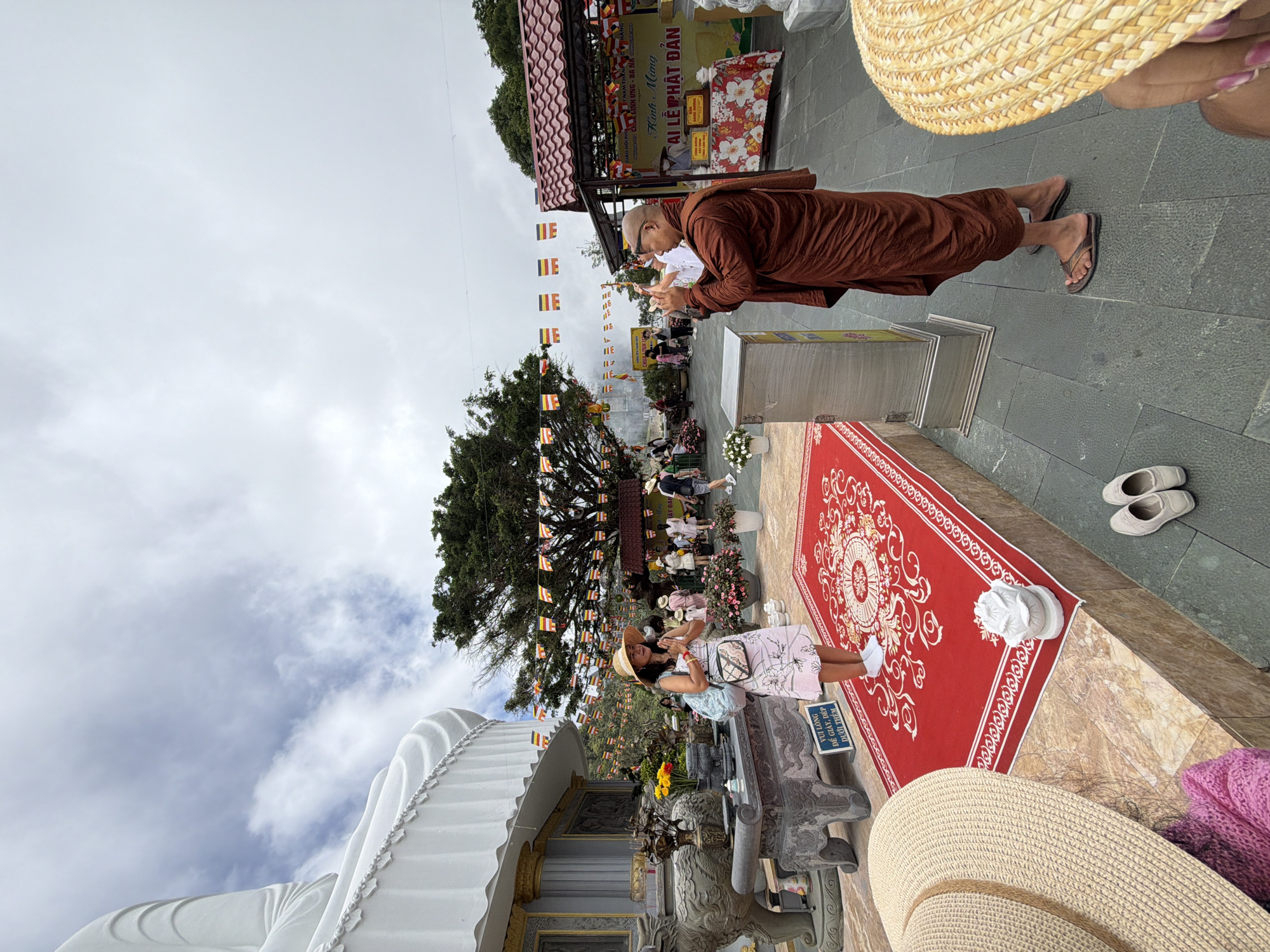 The image depicts a scene outside a Buddhist temple complex in Da Nang, Vietnam, likely on the Son Tra Peninsula, given the presence of a large statue and mountainous terrain. In the foreground, a woman wearing a straw hat and a floral dress stands with her hands pressed together in prayer on a red rug. She is facing a Buddhist monk dressed in a saffron robe, who is holding a cell phone and appears to be taking a photo or video of the woman. Behind the woman, a portion of a large white Buddha statue is visible, along with a stone altar adorned with offerings. A string of colorful Buddhist flags stretches across the scene, contributing to the religious atmosphere. In the background, other visitors are seen exploring the temple grounds, and a table with offerings and religious texts is set up. The sky is overcast, with scattered clouds. The overall environment suggests a tourist destination with religious significance.