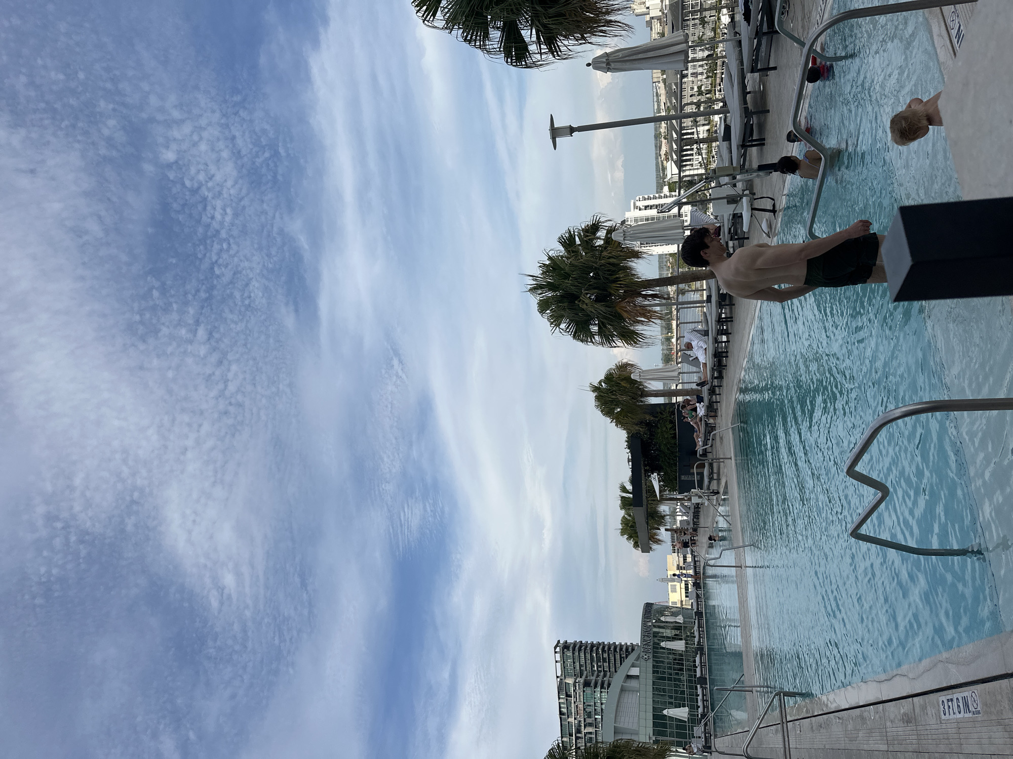 The image captures an outdoor swimming pool scene in Tampa, Florida. The pool water appears clear and blue, reflecting the sky. Several individuals are present, including a man standing near the edge of the pool with his back to the camera, wearing dark shorts. Other people are swimming in the pool and sunbathing on lounge chairs nearby. Palm trees provide some shade along the pool deck. The pool is part of a high-rise building. The sky shows broken clouds with a blue hue. The pool has metal handrails and a concrete deck. In the background, other city buildings are visible.
