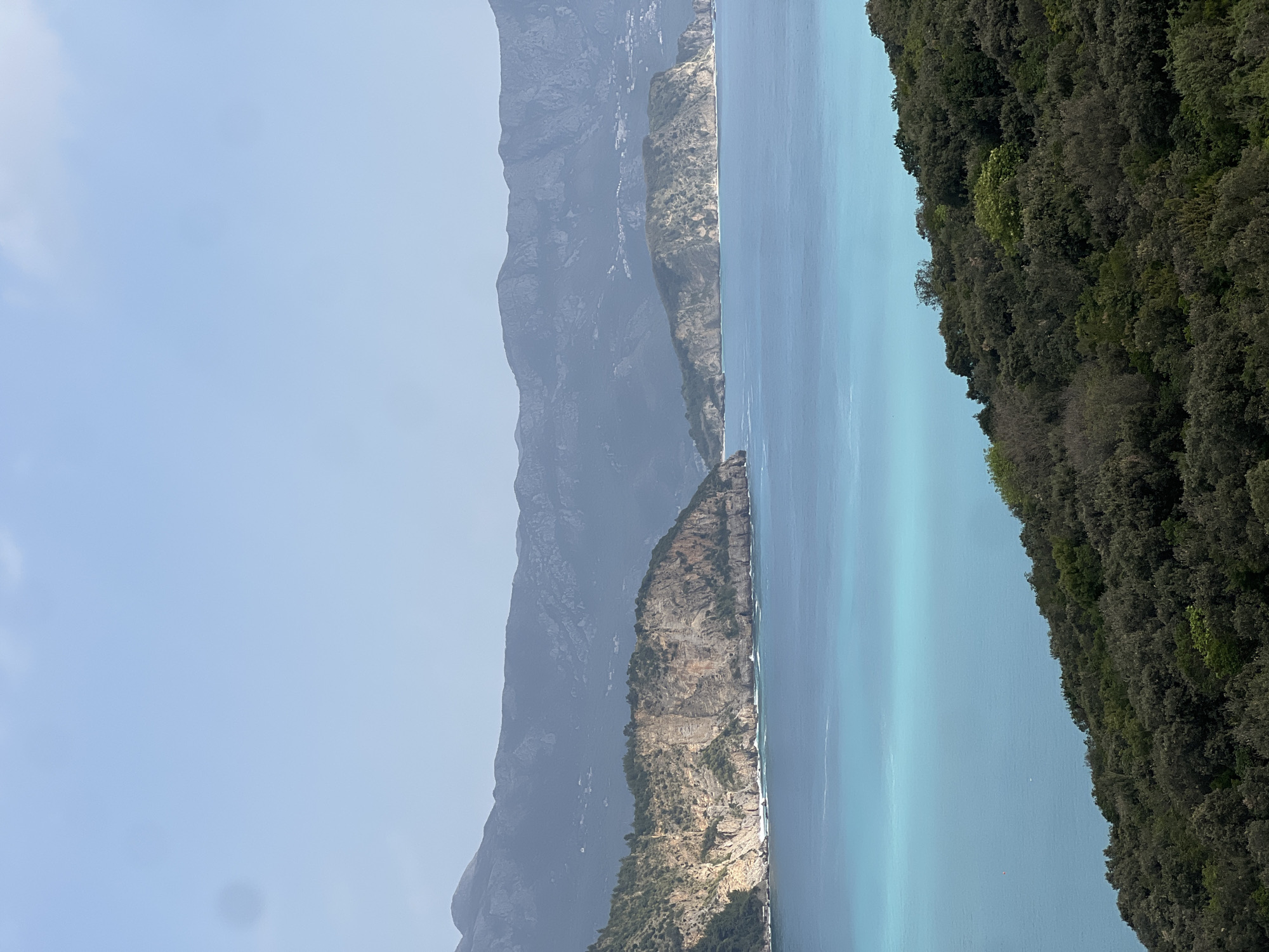 The image depicts a coastal scene in Visnjeva, Montenegro. A body of turquoise water separates a densely forested shoreline on the right from a rugged, mountainous terrain on the left. The mountains fade into a hazy blue distance. The sky is clear and light blue. There are no people or discernible objects present in the view.