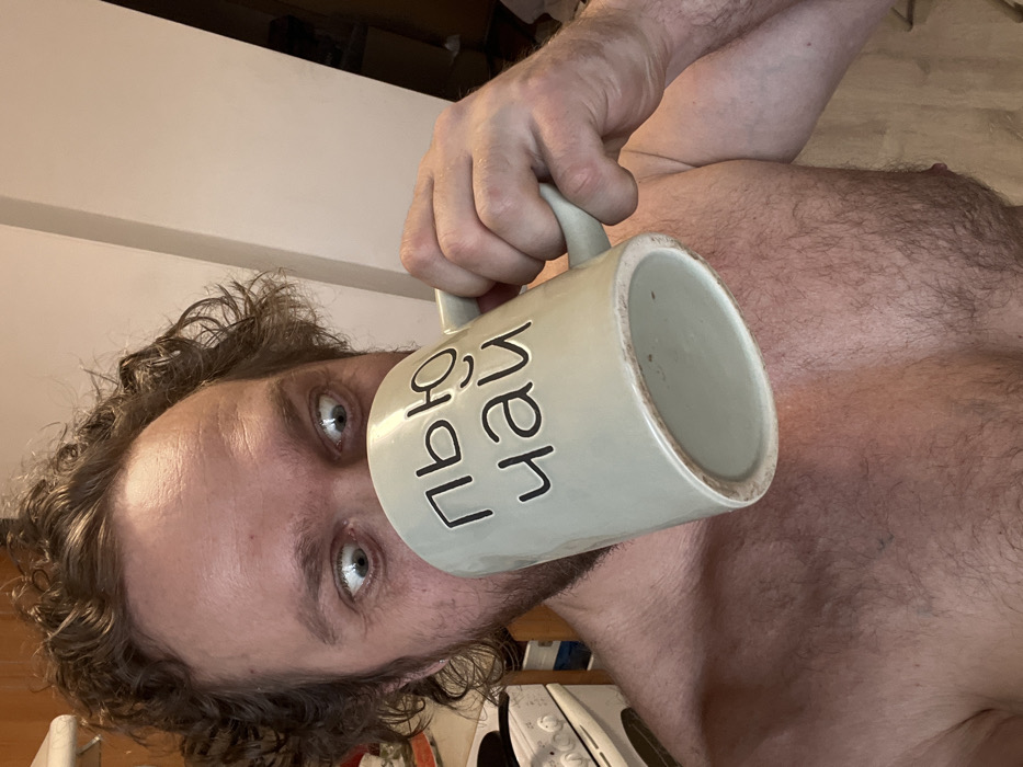 An adult male is depicted in a close-up, chest-up shot within an indoor, domestic setting. The man has curly brown hair and significant body hair on his chest. He appears shirtless. His eyes are wide open, with visible sclera above and below the irises, fixed on the viewer. He is holding a large, light grey ceramic mug to his mouth with his right hand, tilted upwards as if actively drinking. The mug features dark Cyrillic lettering, clearly displaying "ПЬЮ ЧАЙ", which translates to "I drink tea". In the blurred background to the left, a white kitchen appliance, likely a stove or oven, with a black grate is partially visible. The immediate background behind the man consists of light-colored walls. No specific location within the city of Saint Petersburg, Russia, is discernible from the visual information provided; the environment appears to be a private residence.
