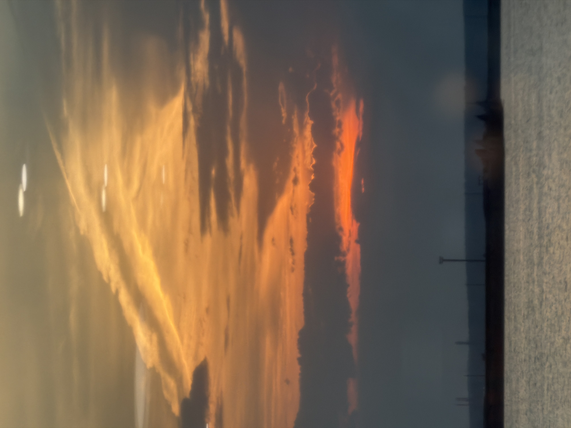 This is a wide, horizontal, slightly blurry photograph of a sunset over a body of water, likely the Kama River, near the city of Chaikovsky, Russia. The sky is dominated by warm hues of orange, yellow, and muted grey clouds. Wispy contrails streak across the upper portion of the sky, catching the sunlight. Several circular, bright white orbs, possibly lens flares or light pollution, are scattered in the upper and middle sections of the sky.
The middle ground of the image features a dark, silhouetted horizon line. On this horizon, several indistinct structures are visible, including what appear to be industrial smokestacks or towers and a tall, slender lamppost. The lower portion of the image is filled with the choppy, grey surface of the water, reflecting the muted light of the sunset. The overall impression is one of a calm, industrial sunset. There are no discernible people or animals in the scene. The scene appears to be devoid of any activity.
