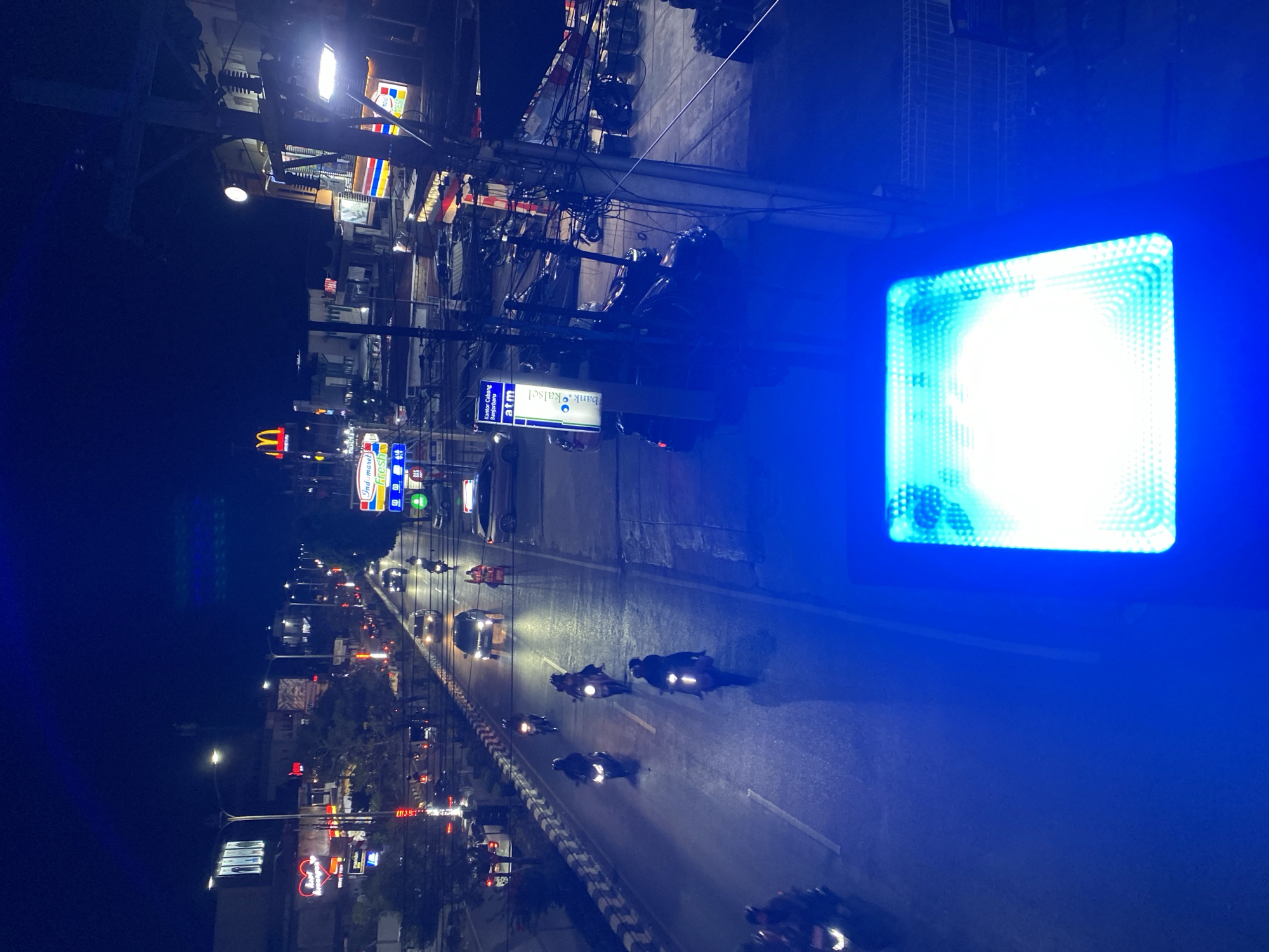 This is a night-time, high-angle shot of a street in Banjarbaru, Indonesia, likely a commercial area. A road with multiple lanes is visible, with cars and numerous motorcycles navigating it. The street is illuminated by streetlights and the glowing neon signs of businesses, creating a vibrant urban night scene. Signs for McDonald's, Indomaret, and a bank ATM are identifiable. Several motorcycles are being ridden by individuals wearing helmets and dark clothing. The foreground is dominated by a bright, blue-lit object, possibly part of the infrastructure or a light fixture. The overall impression is of a busy street at night with ongoing traffic and commercial activity.