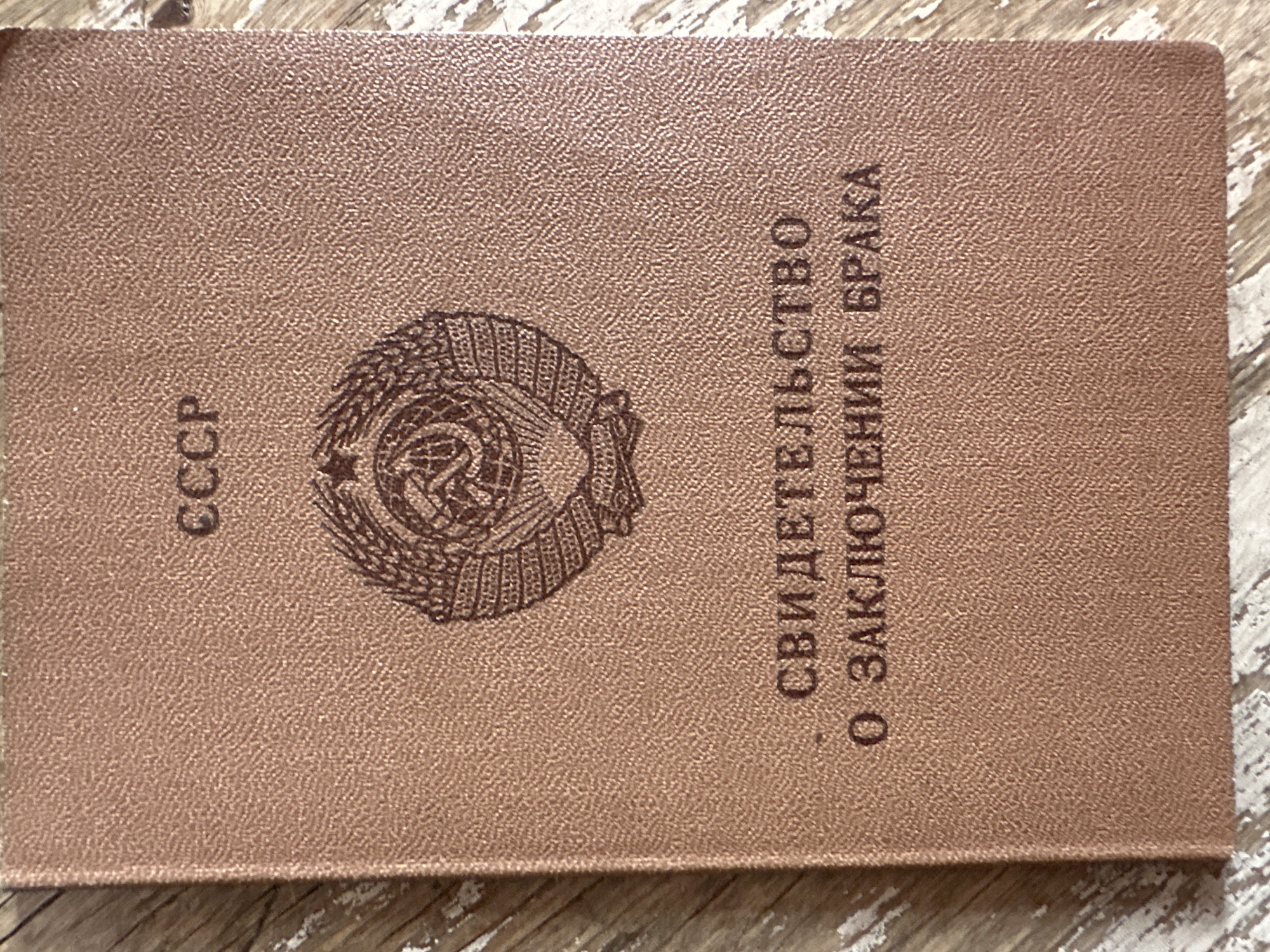 The image shows the front cover of a Soviet-era marriage certificate. The cover is a textured brown material. At the top, in capital letters, is "СССР" (USSR). Below that is the Soviet coat of arms, featuring a hammer and sickle, a globe, and wheat stalks. Underneath the coat of arms, in larger letters, are the words "СВИДЕТЕЛЬСТВО" (Certificate) on the top line and "О ЗАКЛЮЧЕНИИ БРАКА" (Of Marriage) on the bottom line. The document is placed on a surface that appears to be a wooden table with a distressed, white-washed finish. There are no people visible in the image. The image does not contain any information indicating its location in the city of Biysk, Russia.