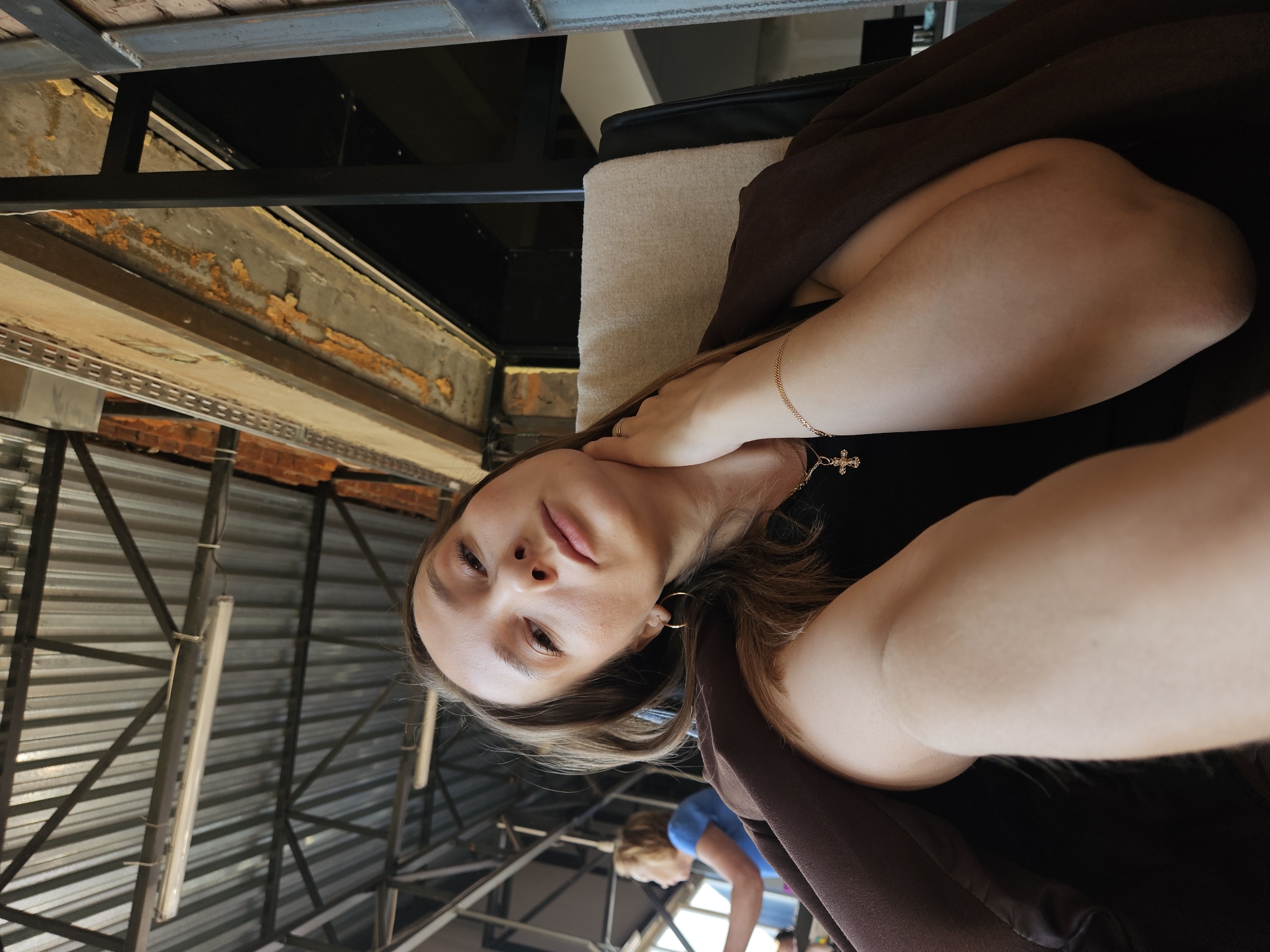 A low-angle selfie of a light-skinned woman with brown hair and gold jewelry (bracelet and hoop earrings) is the main subject. She is wearing a black shirt and brown jacket. Behind her, the interior of a building is visible. The ceiling is made of a metal framework, and there is a woman with blonde hair wearing a blue shirt visible in the background. In the upper left corner, there is a partially visible brick and concrete structure. This photo was taken in the city of Chaikovsky, Russia.