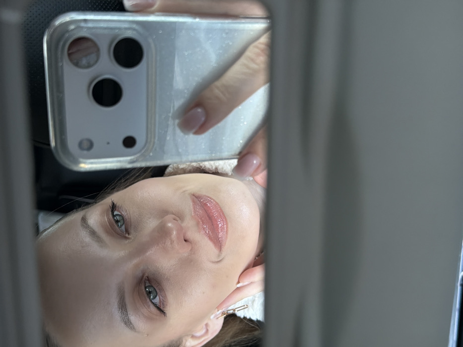 The image presents a close-up, matter-of-fact mirror reflection of a fair-skinned female subject, appearing to be in her twenties or thirties. She has light brown hair, green or hazel eyes with subtle eyeliner, and full lips coated with a glossy, light pink or nude product, conveying a slight smile. She wears a light-colored, textured top, possibly sherpa or fleece, and a gold-colored, bar-shaped dangle earring is visible on her left ear. Her right hand, featuring neatly manicured nails with light pink polish, holds a smartphone, likely an iPhone due to the visible multi-lens camera array, encased in a clear, glittery cover. Her left hand gently touches her jawline. The interaction captured is a selfie being taken via the mirror. The immediate environment is the interior of a vehicle, indicated by the grey overhead panel visible in the lower portion of the reflection and the indistinct, dark background suggesting an enclosed space. No specific geographical landmarks or external identifiers pertaining to the city of Selevino, Russia, are discernible from the visual content.