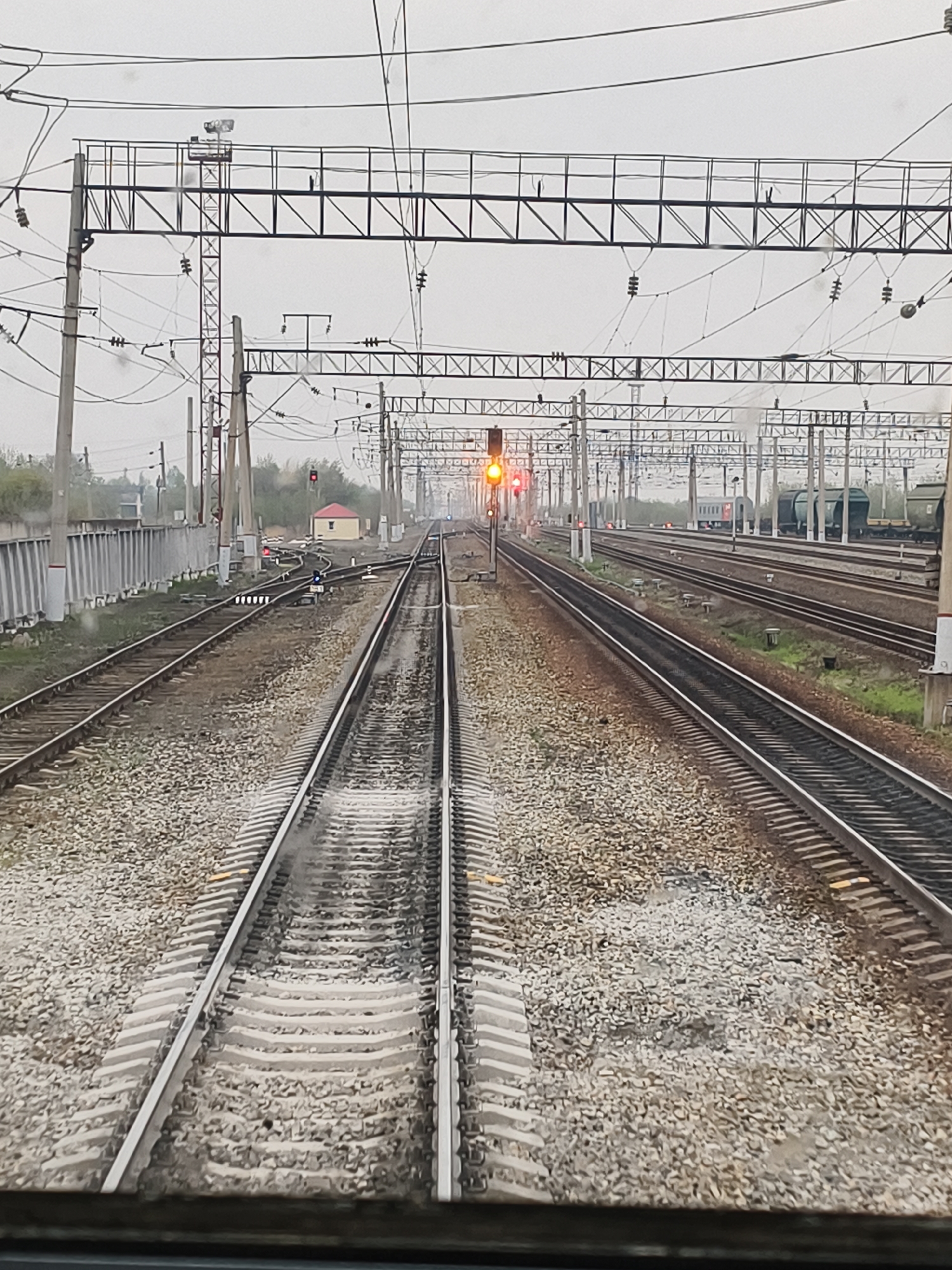 The image shows a view from a train cabin looking out onto a railway yard in an unidentified location. Parallel railroad tracks stretch into the distance, covered in gravel. Overhead, a network of electrical lines and metal support structures is visible. A red traffic signal is lit in the distance. Several train cars are parked on parallel tracks. A small building is to the side, next to the tracks. The day appears overcast.