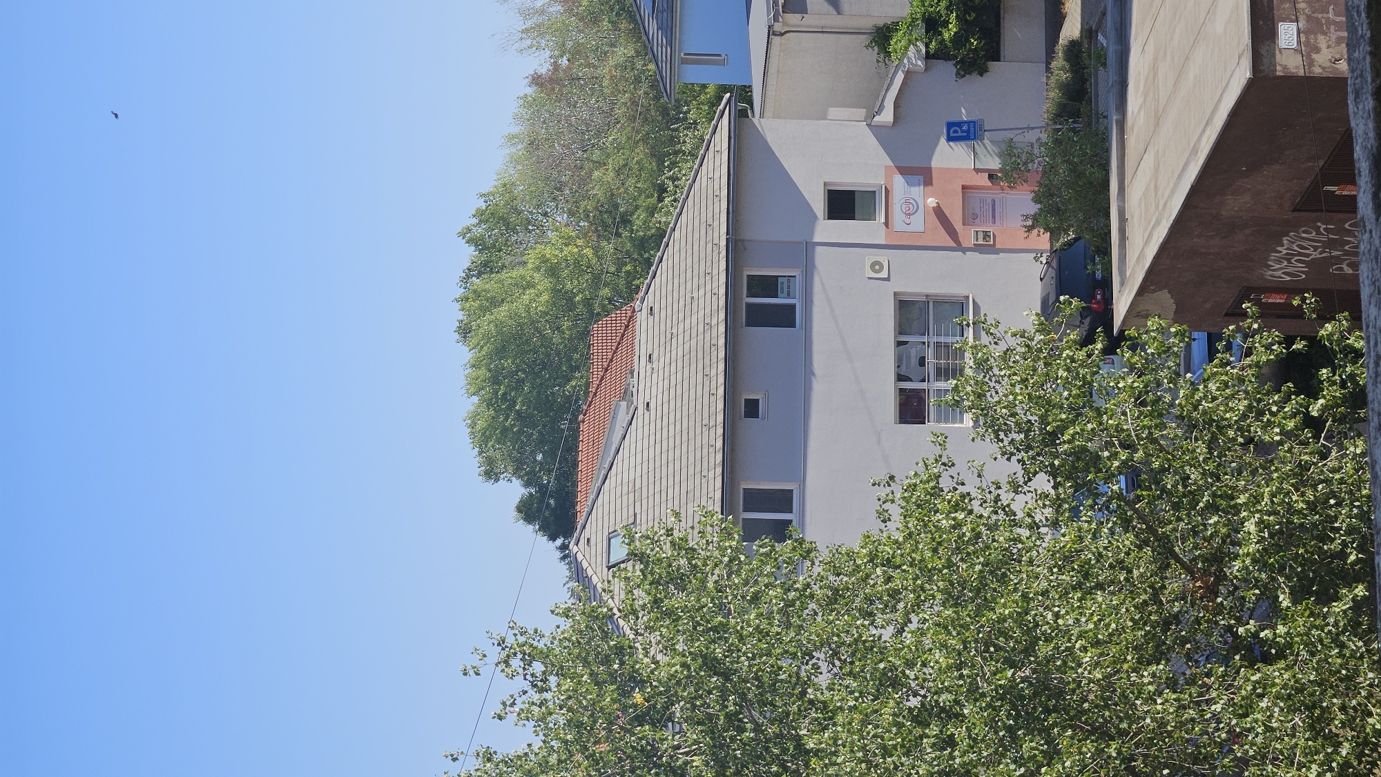 The image shows a three-story residential building with a light gray facade and a gray tiled roof. The building is situated in Bratislava, Slovakia, on a clear, sunny day. A lush green tree with vibrant leaves partially obstructs the view of the lower floors, while the upper floors are visible against a bright blue sky. The building has several windows, some with white frames. The ground floor features a storefront or office space with a pink accent wall and a rainbow logo. A blue parking sign with a white 'P' is visible near the entrance. Several cars are parked in front of the building, including a dark-colored SUV. The surrounding environment is characterized by more greenery and other buildings in the background, one of which is painted light blue. A small bird is visible in the sky.