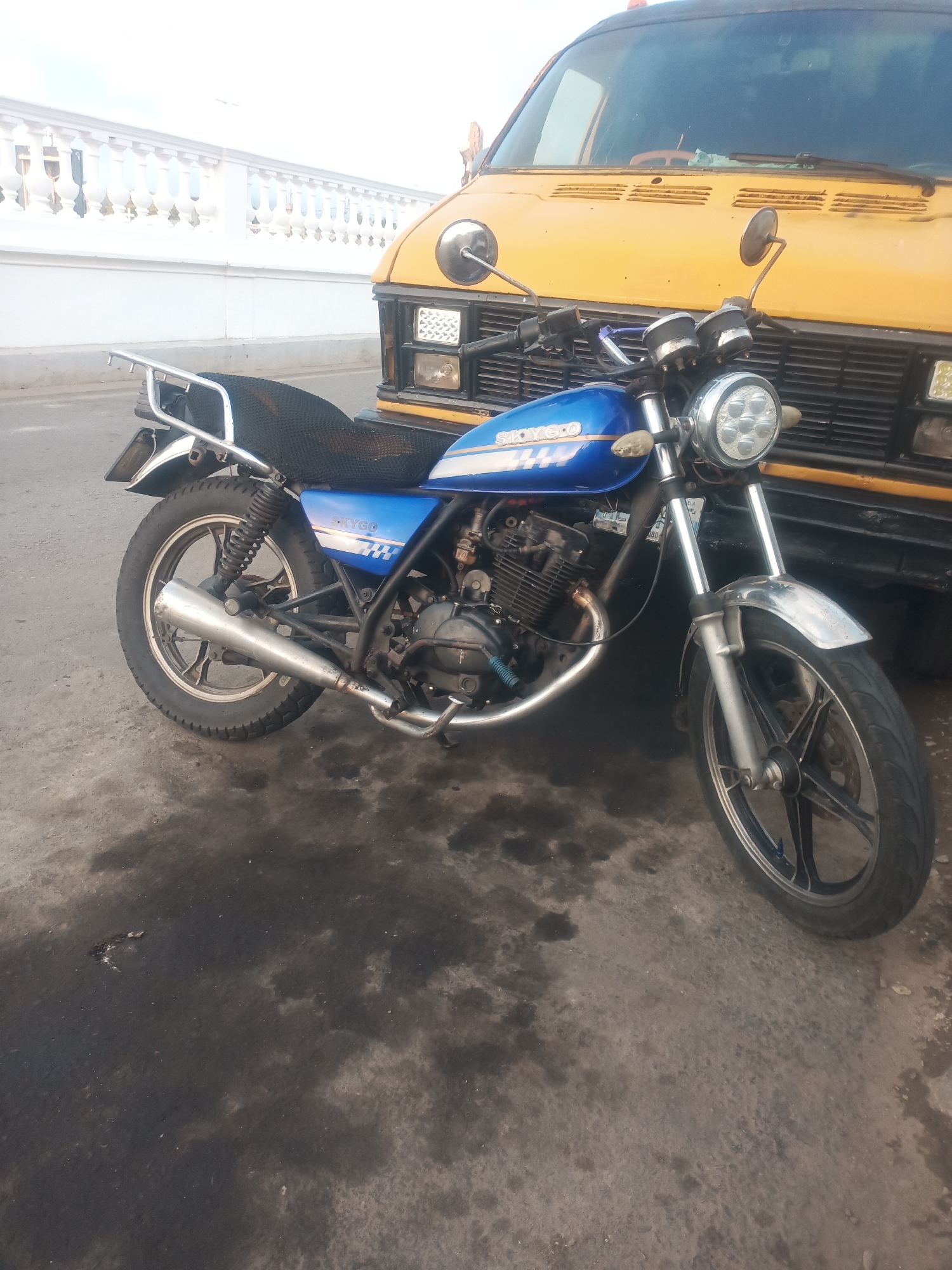 In Juan Griego, Venezuela, a blue and white motorcycle is parked on an asphalt surface next to a yellow van. The motorcycle has a chrome exhaust pipe and a black seat. The van is parked closely behind the motorcycle. A white decorative railing is visible in the background, suggesting the scene is near the coast or a promenade. The lighting suggests it is daytime, possibly overcast due to the brightness of the white railing and the van. The ground is stained with dark patches, possibly oil or water. There are no people visible in the image.