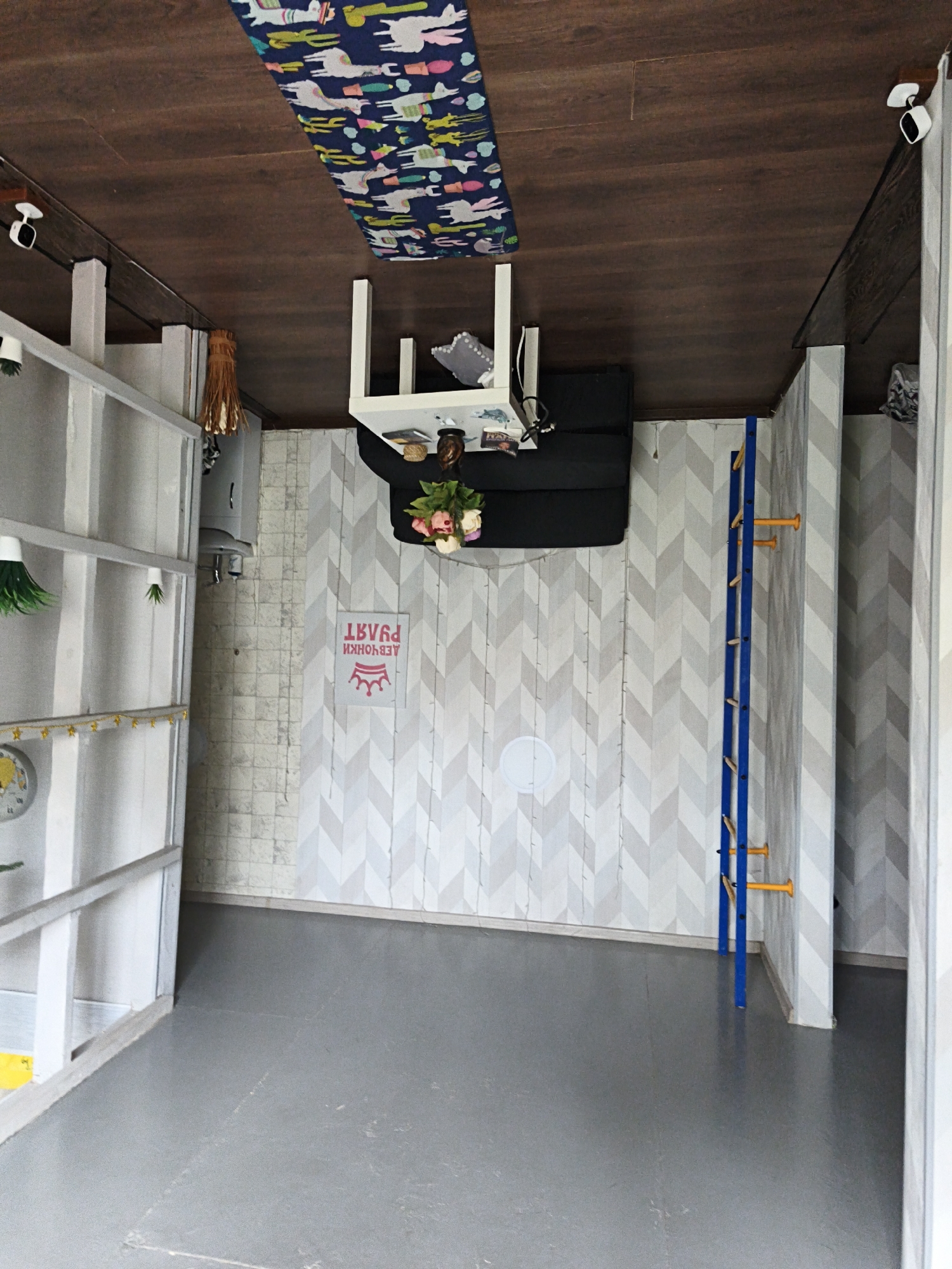 This image captures an interior room designed as an optical illusion, where the furniture appears to defy gravity, giving the impression of an upside-down space. The photo itself is rotated 90 degrees counter-clockwise.
The primary subjects are the room's decor and furnishings. A dark wooden ceiling, in reality the floor in the image's orientation, features a vibrant blue rug adorned with llamas, cacti, and other playful shapes. Two white security cameras are mounted to beams. Attached to what would normally be the ceiling, but is presented as the floor in this illusion, is a white table with a black base and a black sofa underneath. A bouquet of pink and white flowers, books, and other decorative items are on the table, while a cushion rests on the sofa.
The walls are distinctly decorated. The left wall features a tall, white shelving unit holding various green decorative items, a clock with a star garland, and other small objects. A woven light fixture hangs from a beam. The back wall is covered with a light gray and white chevron patterned wallpaper, with a section of white brick-patterned wallpaper below it. A prominent red sign with a crown logo and the Cyrillic text "ДЕВЧОНКИ ПРАВЯТ" (translating to "Girls Rule") is displayed on this wall. A blue wall-mounted ladder with yellow rungs, possibly for exercise or play, is affixed to the right side of the back wall. The right wall continues the gray and white chevron pattern. The actual floor of the room is a plain, dark gray surface.
No people are visible. The room's setup suggests it is a novelty attraction or an exhibit designed for visitors to experience and photograph the illusion of an inverted environment. The lighting is even, typical of an indoor setting. This location is contextually identified as Sevastopol, Ukraine.