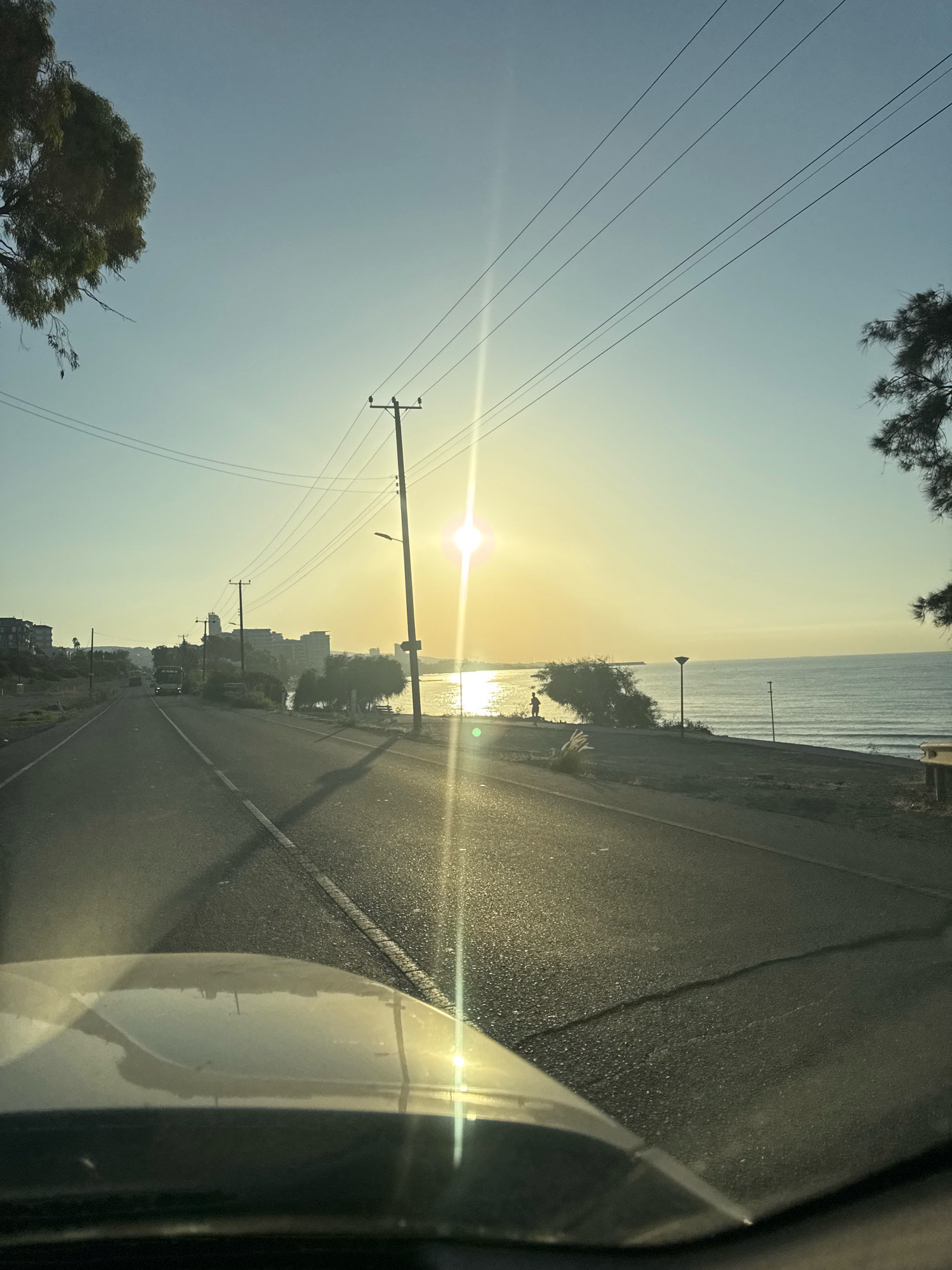 The image depicts a coastal road scene in Agios Tychona Community, Cyprus, during what appears to be sunset or sunrise. A paved road runs diagonally through the frame, leading towards a body of water. The road is lined with power lines and poles on the right side. The sun, a bright starburst, is positioned slightly to the right of the center, casting a strong lens flare and illuminating the water. On the right side of the road, there's a shoreline with some vegetation and a few people visible near the water. A distant cityscape with buildings is visible on the left side of the road, further inland. The sky is clear with a gradient of light blue to pale yellow. The foreground shows the hood of a vehicle, suggesting the photo was taken from inside a car.