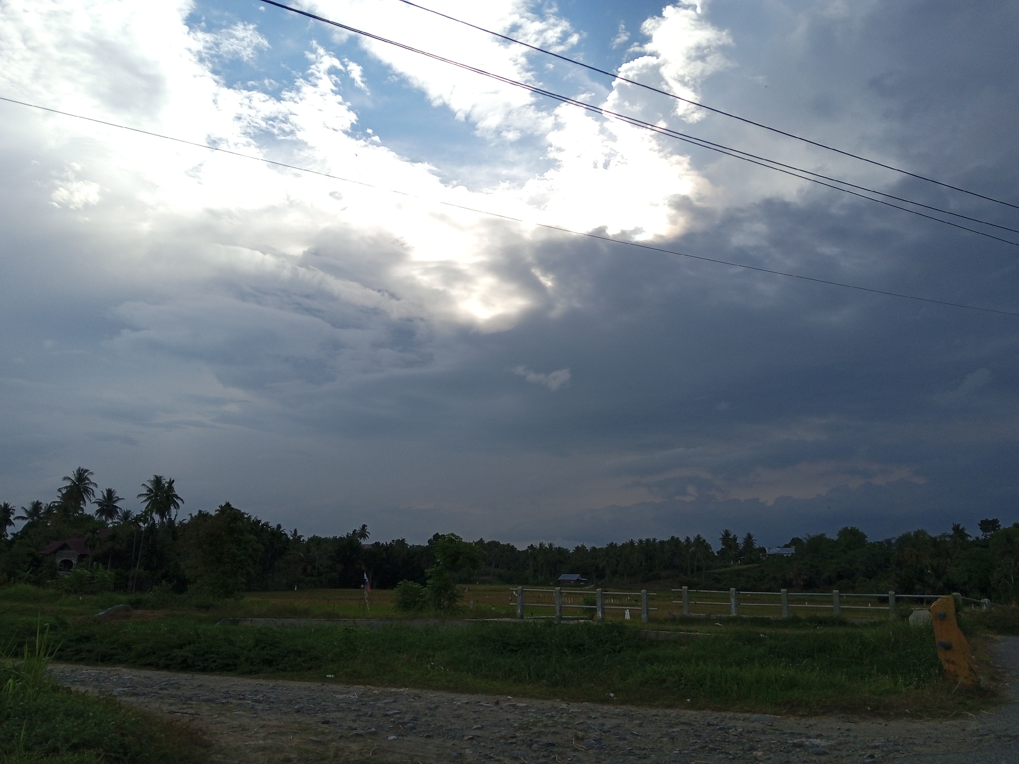 The image presents a view of a rural landscape under a dramatic sky. The sky is filled with a mix of white and dark gray clouds, with sunlight peeking through a gap in the cloud cover. Power lines run across the upper right portion of the frame. Below the sky, the landscape consists of green foliage and trees. In the middle ground, a light-colored fence stretches across the scene. The foreground features a gravel road. The location appears to be Cotjrat, Indonesia, judging by the tropical vegetation and overall setting. No people are visible in the image.