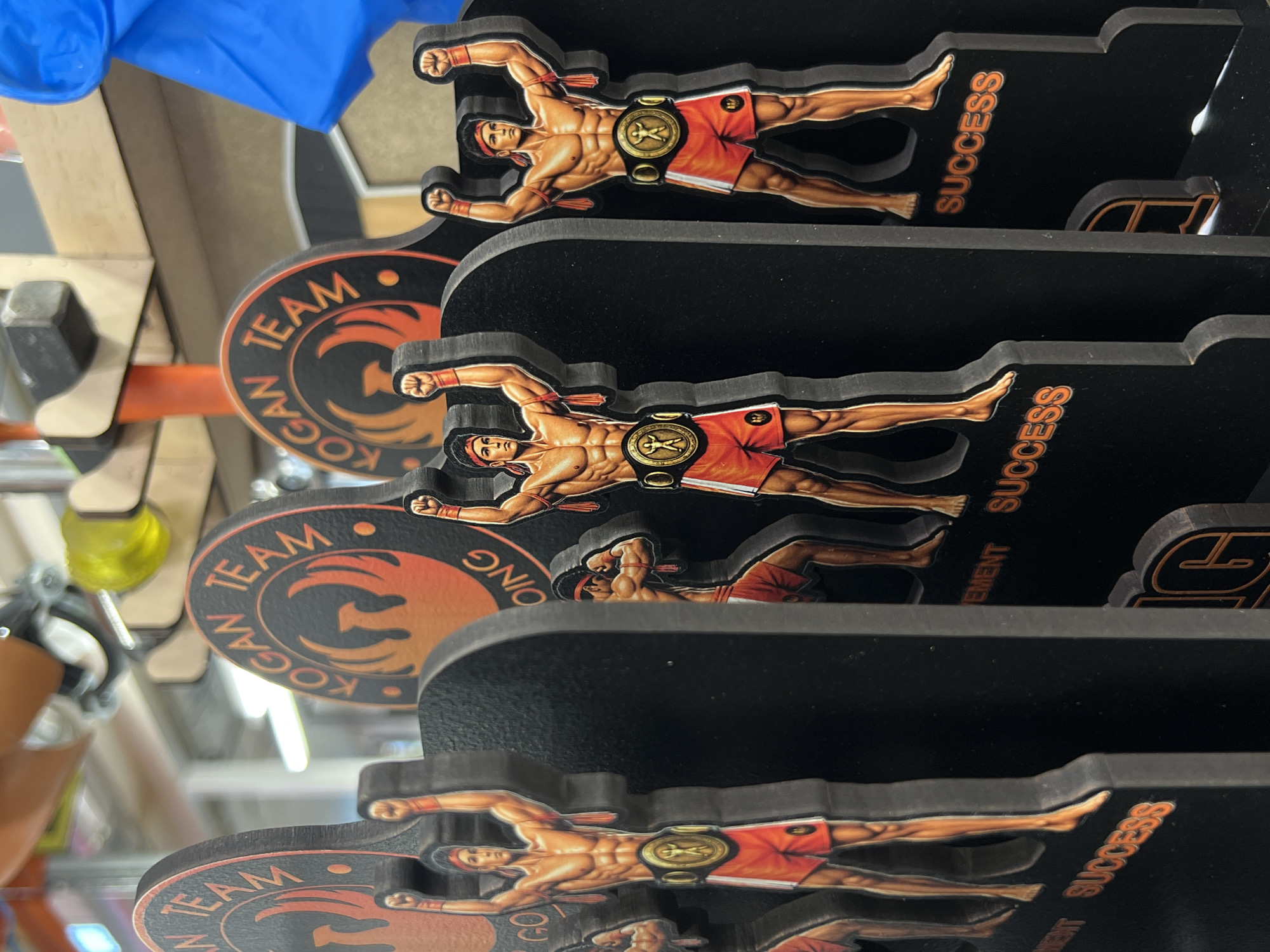 The image displays a close-up, angled view of stacked black display shelves, each featuring a cutout of a muscular, shirtless man wearing red shorts and a championship belt. The figures are depicted in a dynamic, horizontal pose, arms outstretched. On each shelf, there is a circular emblem with orange and black graphics. Text on the emblem reads "TEAM KOGAN". To the right of each figure, printed vertically in orange letters, is the word "SUCCESS". The figures appear to be decorative items or small statues. The background is blurred, but suggests an indoor environment with some shelving and possibly a blue glove visible in the upper left corner. The lighting is bright, with some fluorescent lights potentially visible in the background. The image focuses on the repetition of the figures and the text, suggesting a theme of achievement or martial arts.