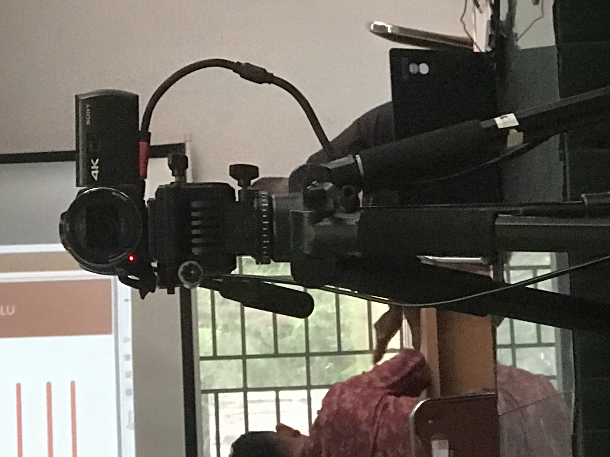A Sony 4K camera is mounted on a black support rig. The camera is positioned near a window with a view of the outside, visible through the window's panes. A person wearing a pink floral shirt is also visible. A portion of a presentation screen with some red vertical bars is visible on the left. The image was taken in Lhokseumawe, Indonesia.