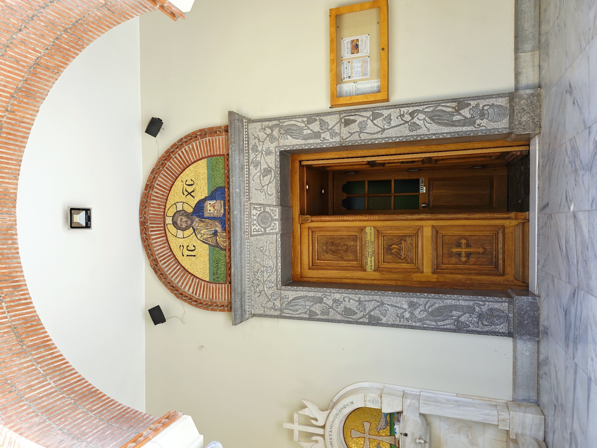 The image shows the entrance to a church, featuring a wooden door with intricate carvings and a mosaic of Jesus Christ above the entrance. The mosaic is set within an arched frame decorated with bricks and stones. To the left of the door, there is a marble font with a cross and a dove mosaic. A bulletin board with notices is visible to the right of the door. The floor is made of marble tiles. The overall scene appears to be from a sunny day, with a bright, well-lit environment. This entrance is located in Agios Nikolaos Municipal Unit, Greece.