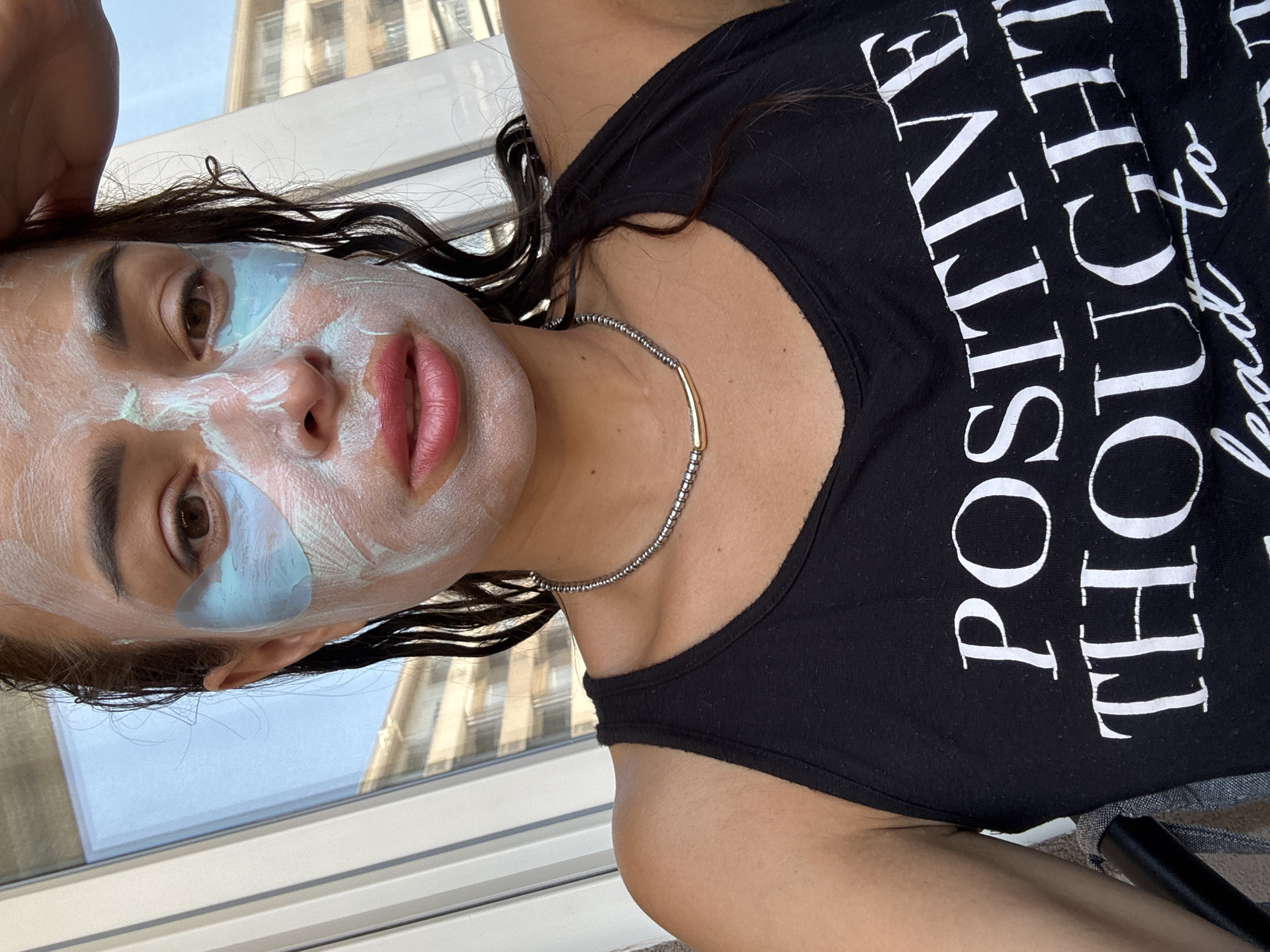 A close-up, waist-up shot shows a young woman with dark hair and brown eyes, her face partially covered with a white and blue face mask, as she applies a mask. She is wearing a black tank top with white text that reads "POSITIVE THOUGHTS LEAD TO". A beaded necklace with a gold accent is visible around her neck. She is indoors, with a window in the background showing a blurry view of buildings. The lighting is bright, suggesting daylight.