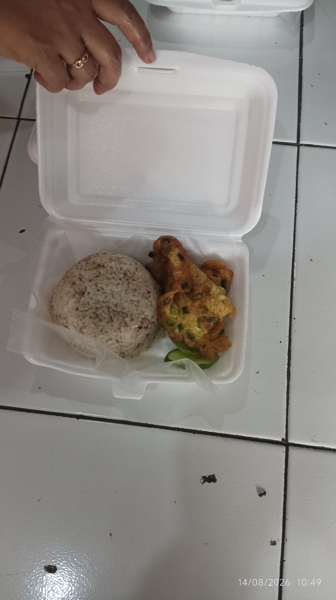 The image shows a Styrofoam food container with food inside. The container is open and placed on a tiled surface. Inside the container, there is a mound of rice and what appears to be fried food, possibly fritters or patties, with some sliced cucumber. A hand wearing a ring is visible in the upper left corner, holding the edge of the container lid. The background shows a tiled floor with some dark marks. A timestamp, "14/08/2026 10:49", is visible in the lower right corner, suggesting the image was taken in Tasikmalaya, Indonesia.