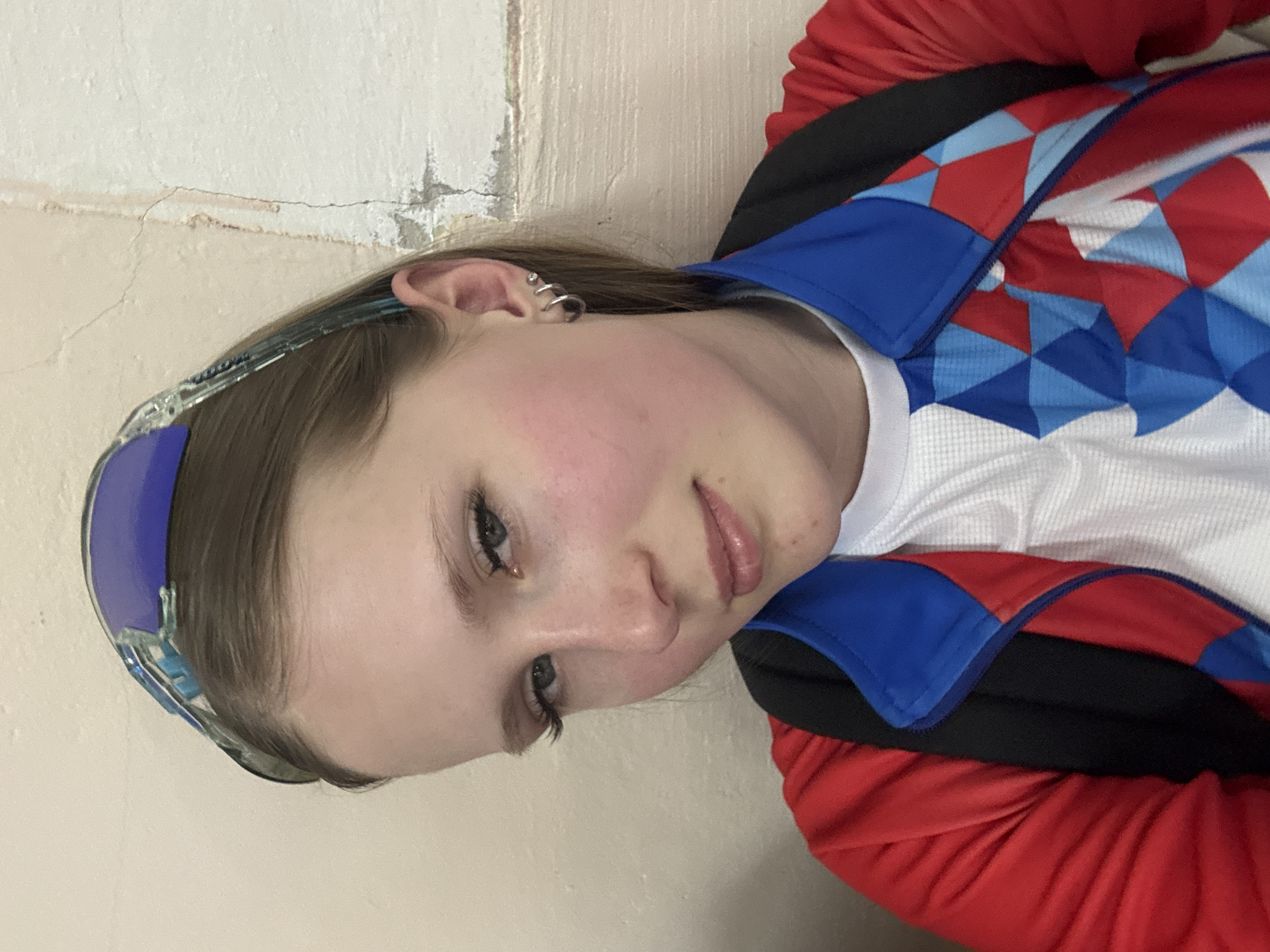 A young woman is pictured indoors, appearing to lean against a light beige wall with visible cracks and imperfections. She wears a pair of blue-tinted eyewear on her head. She has fair skin with slightly flushed cheeks and multiple small hoop earrings on her left ear. She is wearing a patterned athletic jacket; its design includes red, white, and blue diamond shapes on the front. The red jacket is black underneath. Her expression is somewhat neutral. The location is potentially Usolye, Russia, based on provided context.