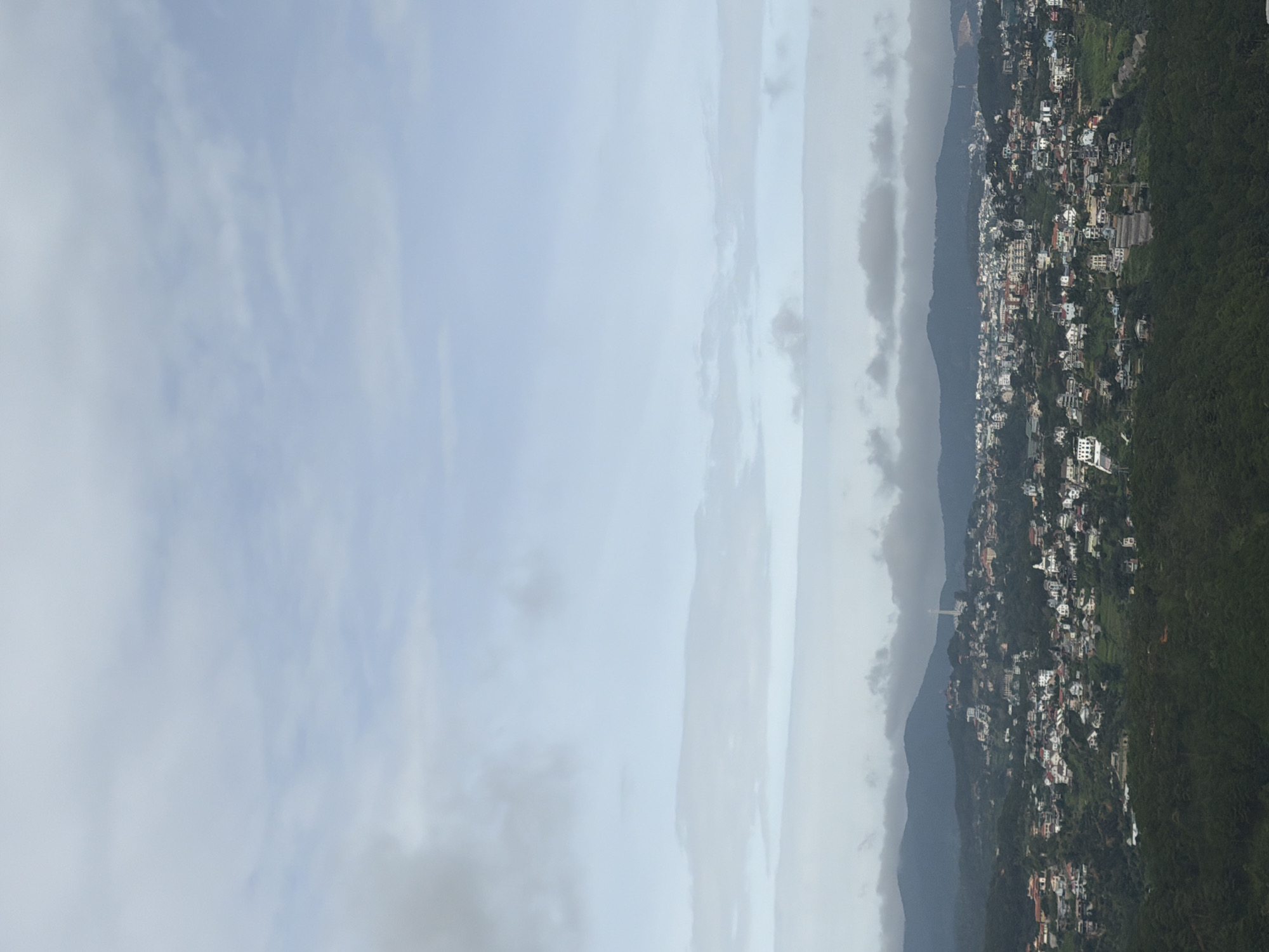 The image shows a panoramic view of a city nestled among rolling hills under a cloudy sky. The city is densely populated with numerous buildings of varying sizes and architectural styles, predominantly white with red roofs. Some larger structures, possibly churches or public buildings, are visible among the residential areas. A tall, slender tower or monument stands prominently on a hilltop in the distance. The hills surrounding the city are covered in lush green vegetation, with trees and foliage creating a natural border. The sky is filled with a mix of light and dark clouds, suggesting overcast weather. The overall impression is one of a vibrant city situated in a scenic, natural landscape. The image does not contain any discernible individuals, actions, or interactions. The specific location of Xuân Thành, Vietnam is inferred from external context and is not explicitly visible within the image itself.